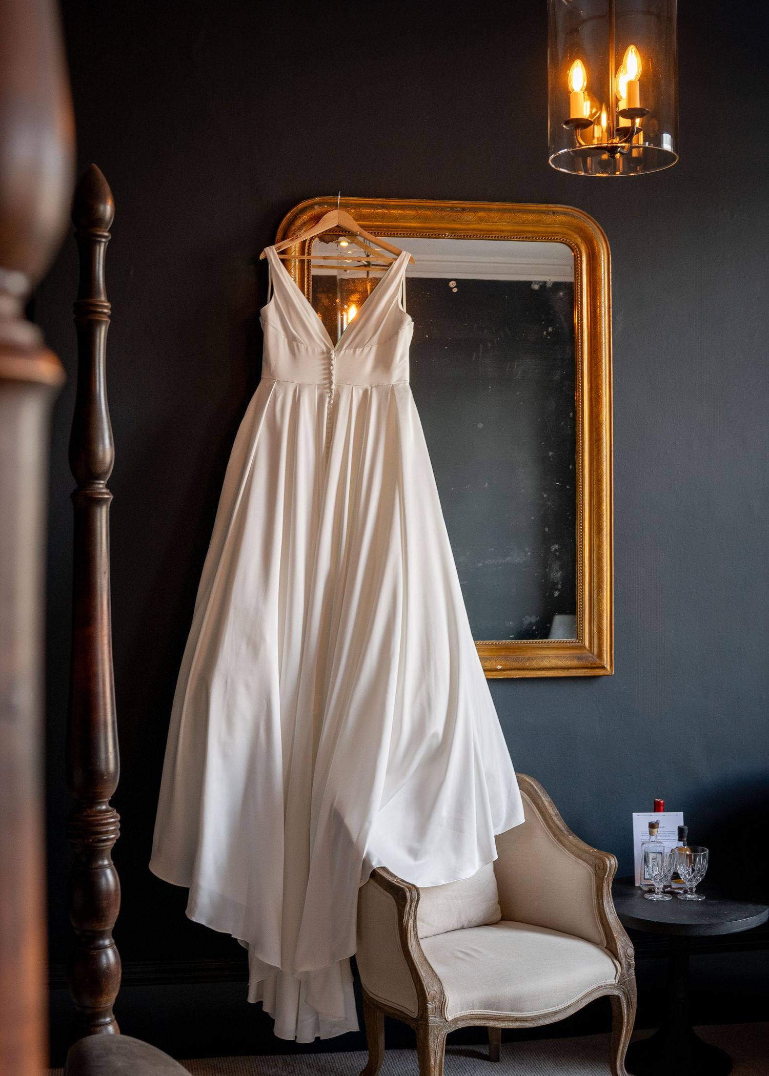 White wedding dress hanging in a dark room with gold-framed mirror and vintage decor.