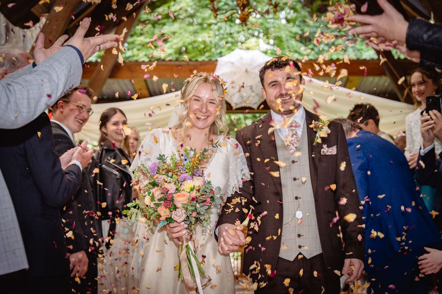 Joyful newlyweds walk through colorful confetti shower at outdoor woodland wedding ceremony exit.