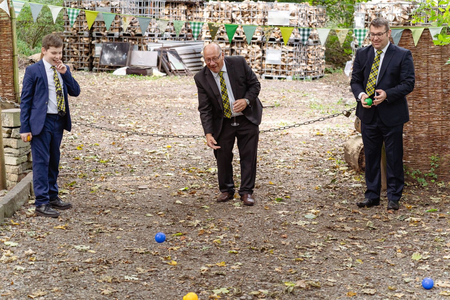 Three men playing boules outdoors, with colorful balls scattered on the ground in a garden setting.