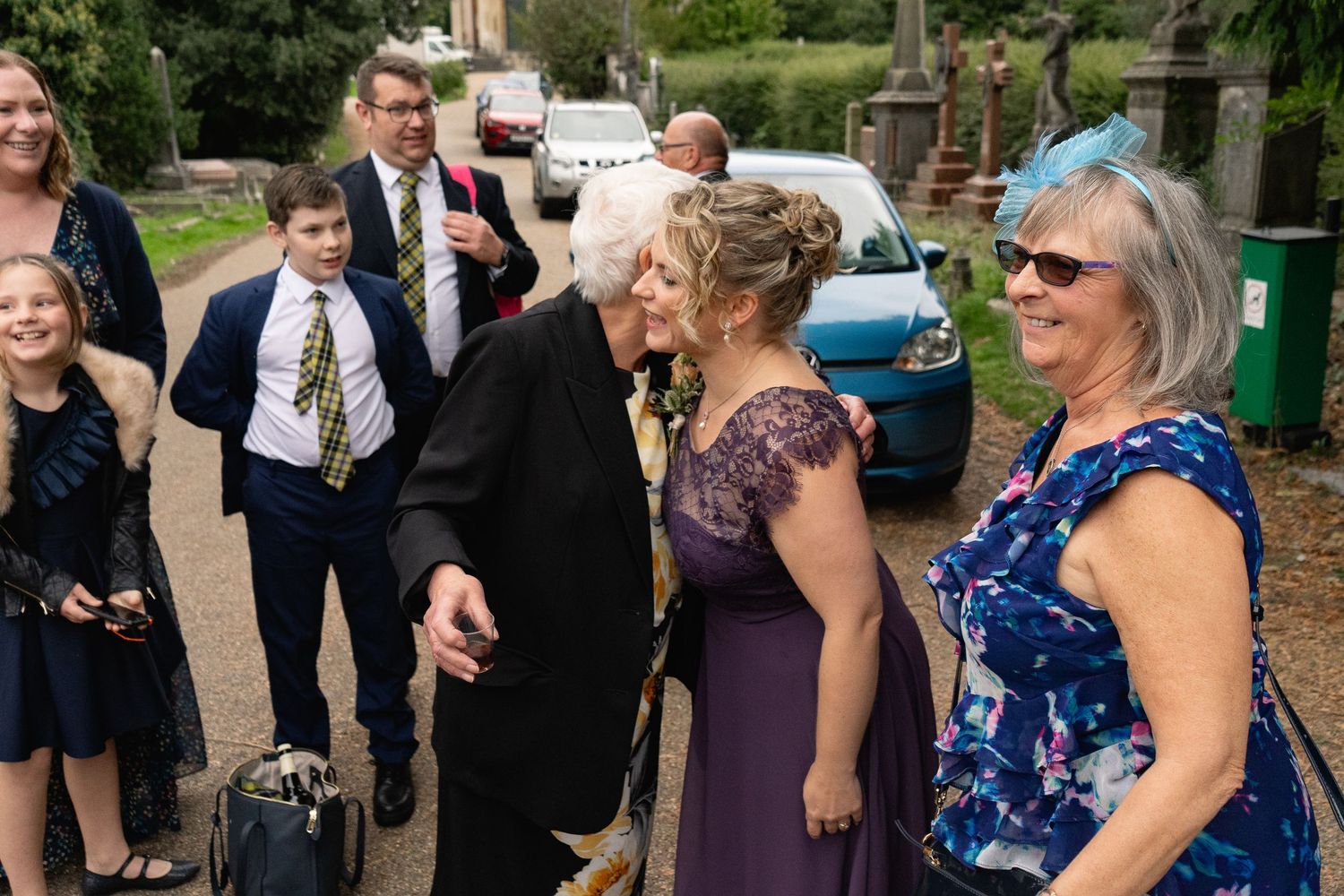 Wedding guests in formal attire greet each other outside a church, with women in purple dresses and a boy in a suit.