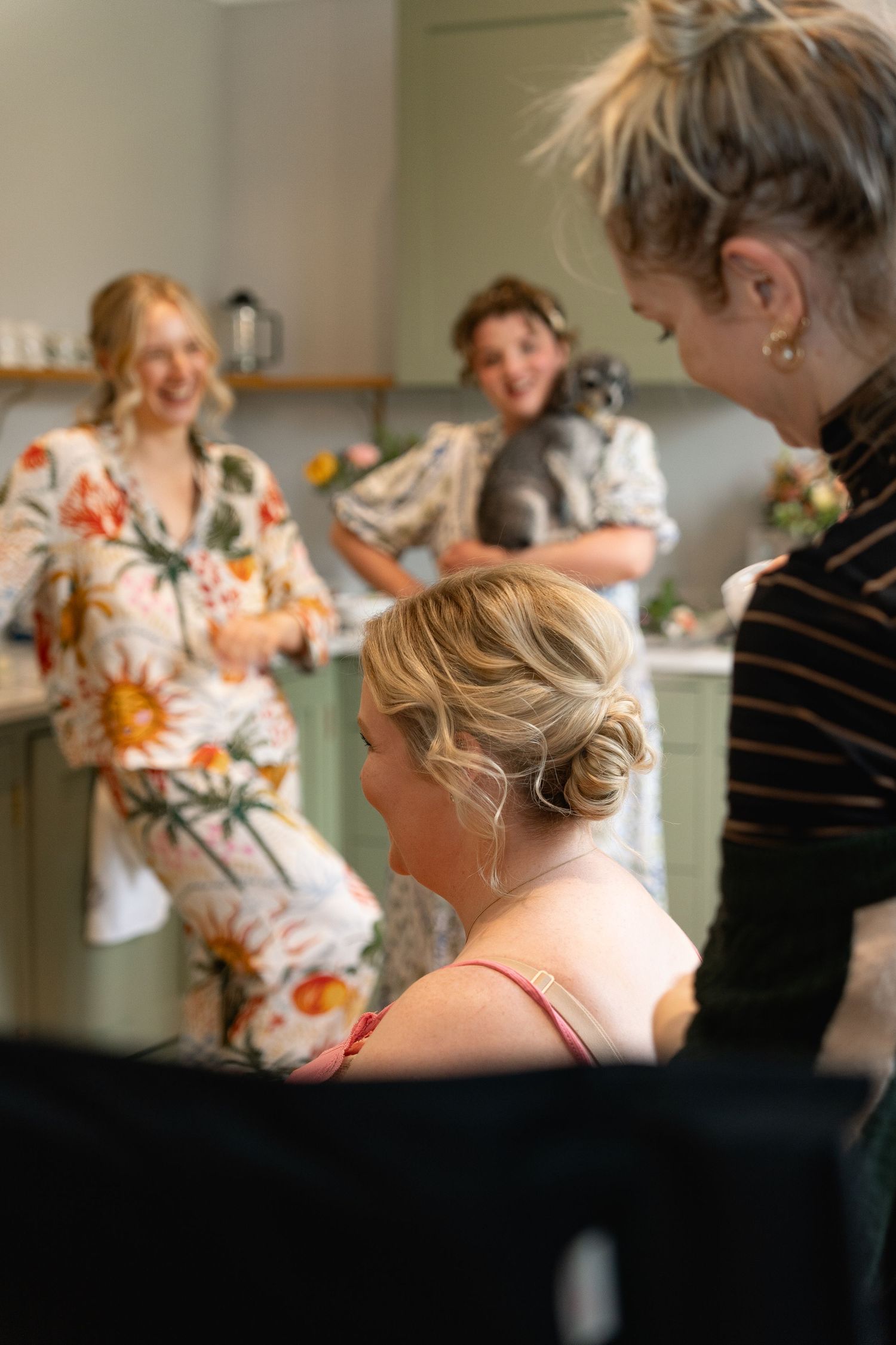 Hair stylist working on blonde bride's updo while bridesmaids laugh in floral pajamas.