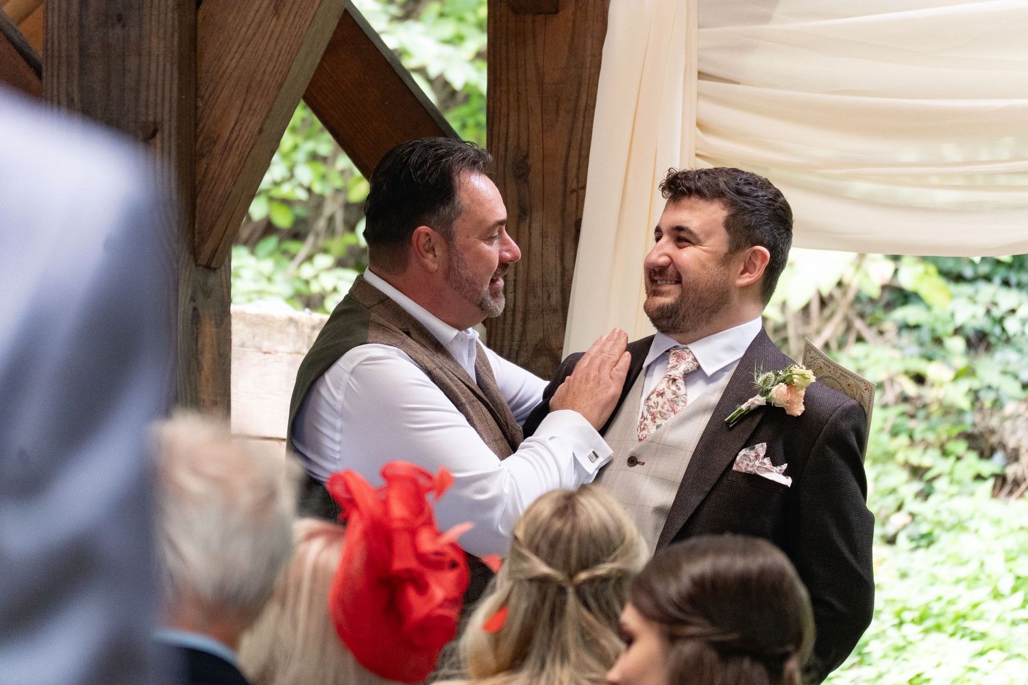 Two grooms share a joyful moment at their outdoor wedding ceremony beneath a wooden arbor.