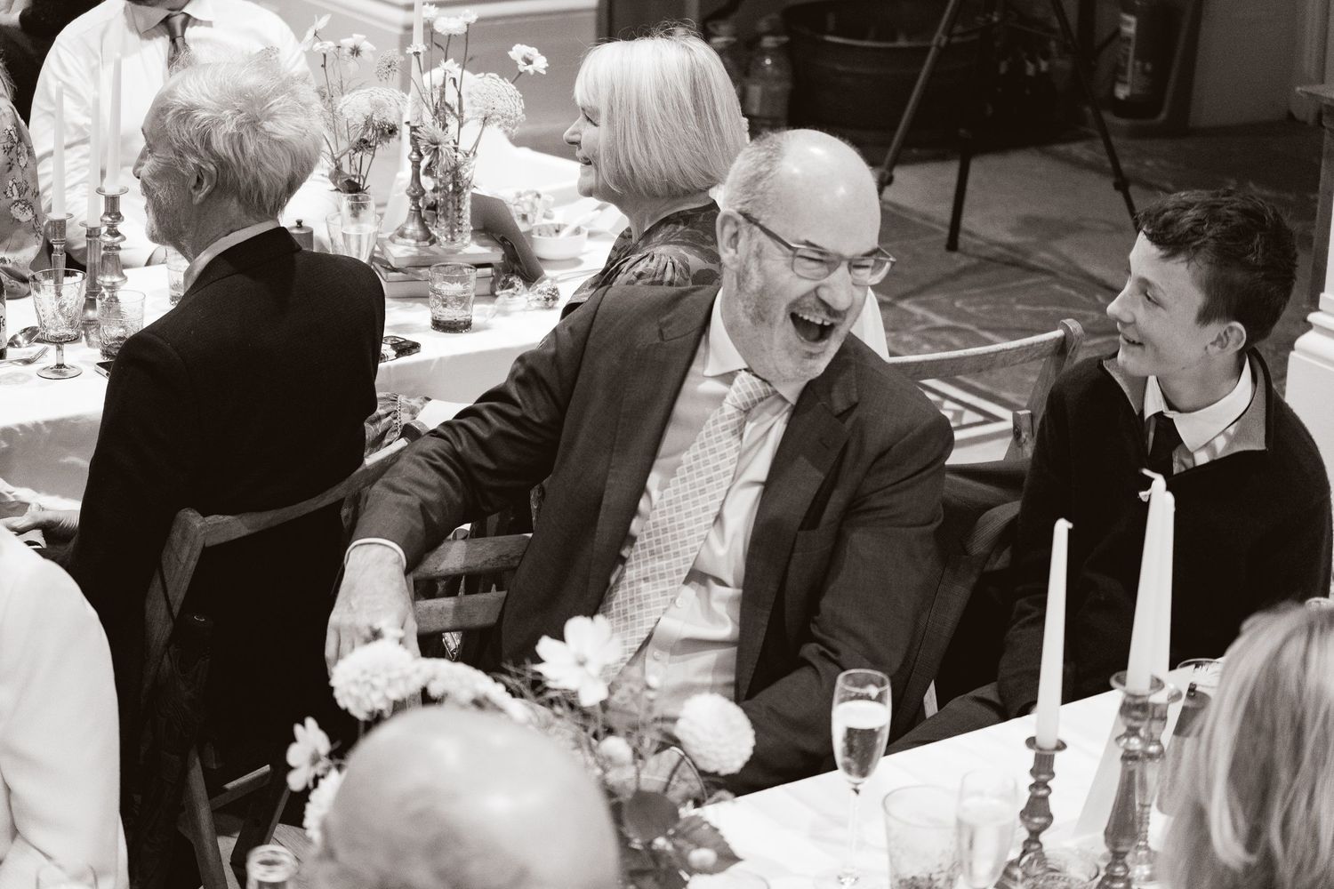 Black and white photo of guests laughing joyfully at a wedding reception dinner table with candles and flowers.