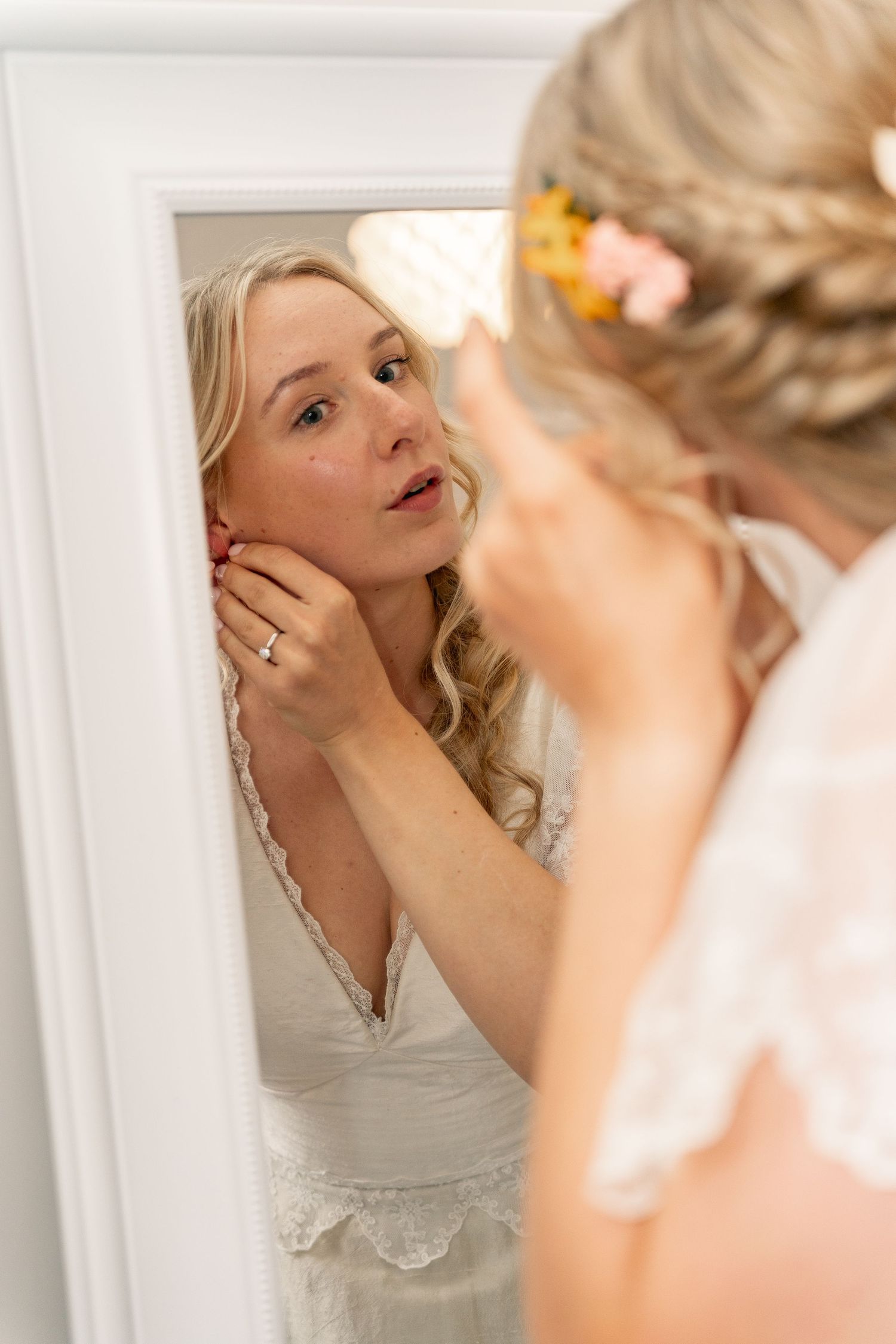 Blonde bride in lace wedding dress puts on earrings while looking in white mirror with floral hair accessory.