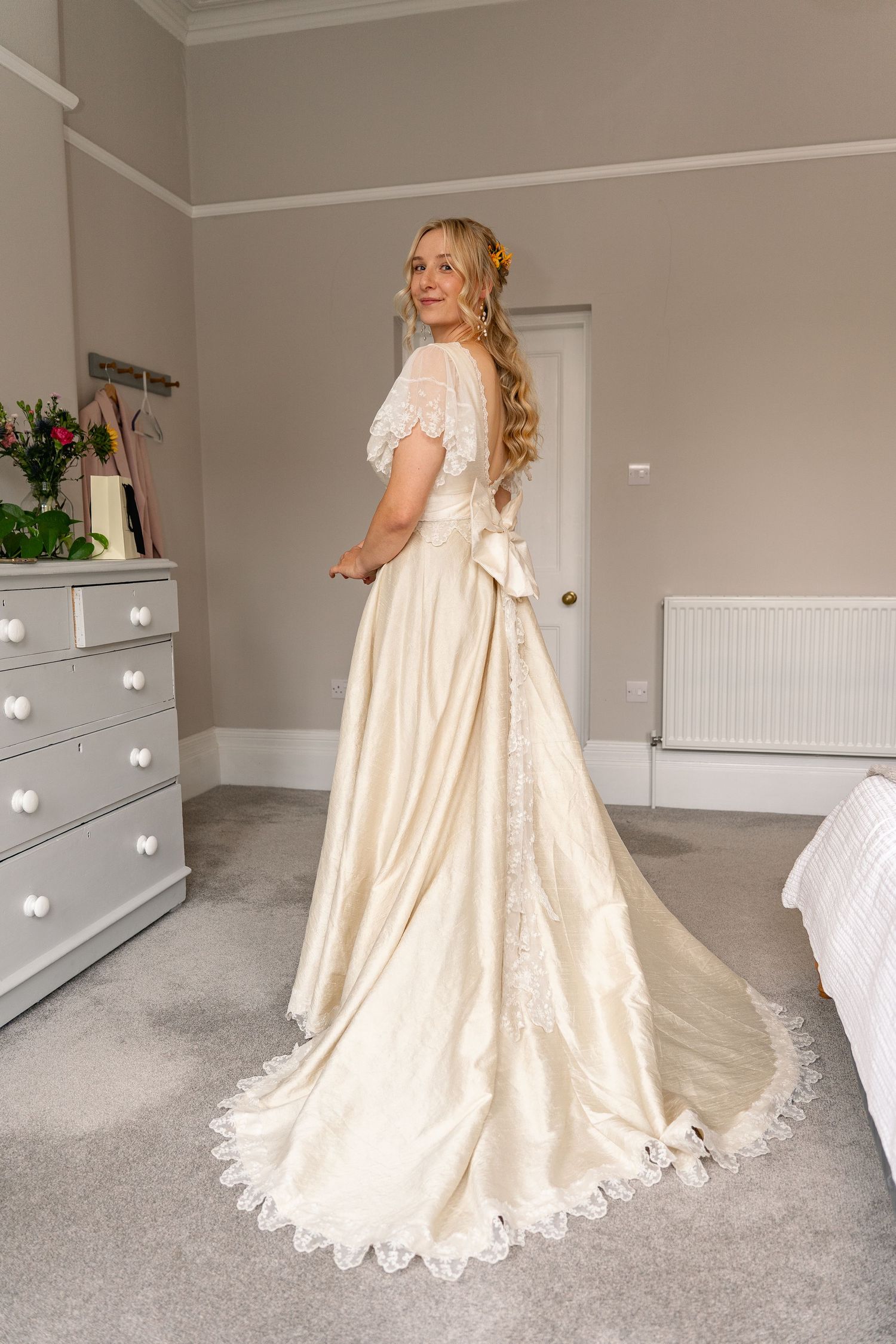 Bride in elegant ivory satin wedding gown with lace trim and bow, posing in a bright bedroom.