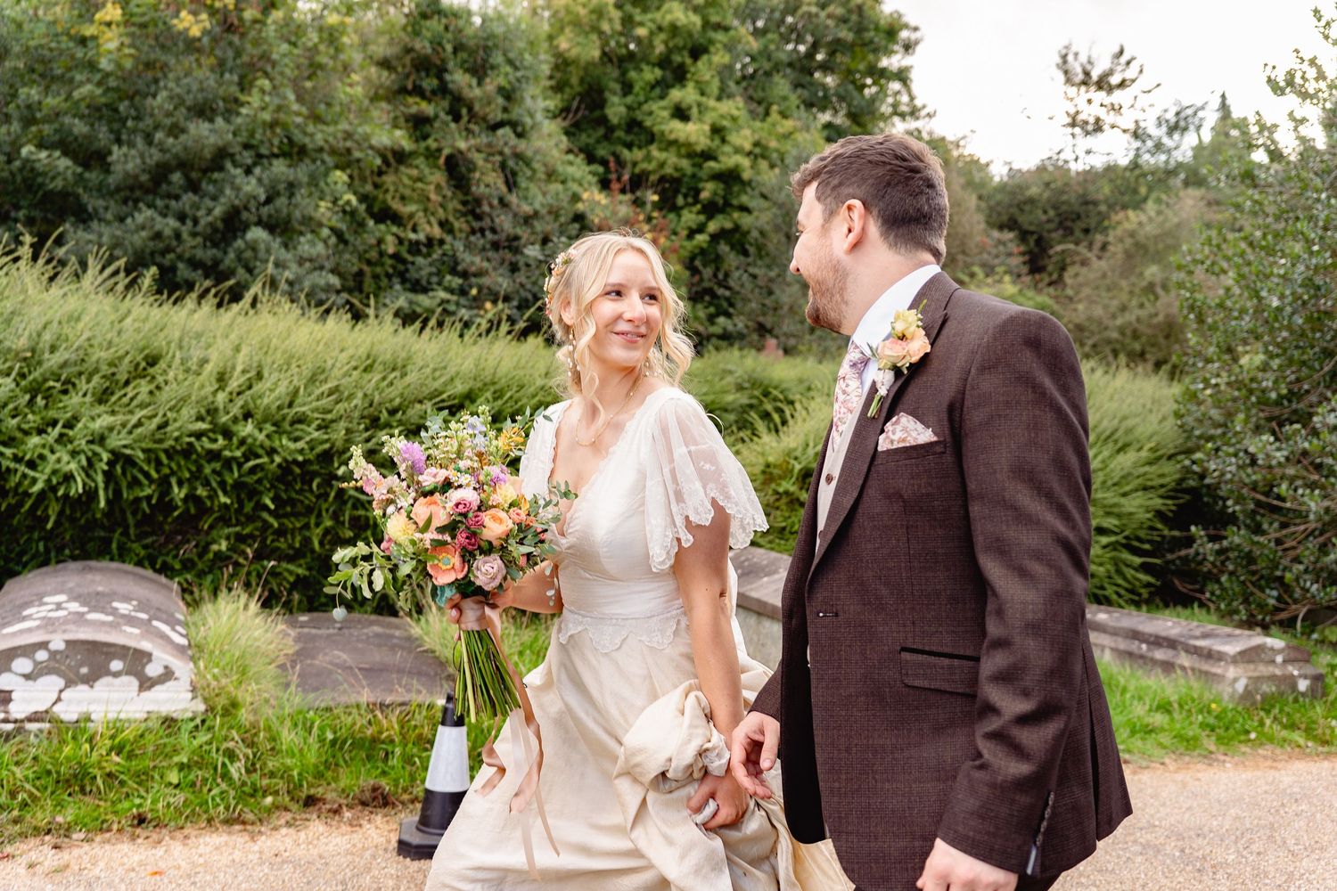 Bride and groom walking hand in hand outdoors, smiling at each other, bride holding a colorful bouquet, groom in brown suit.