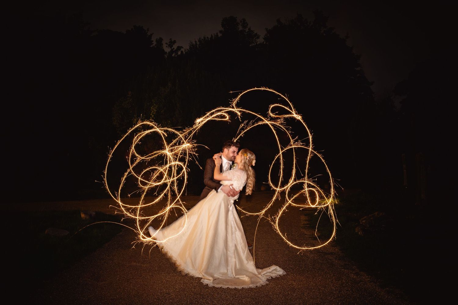 Bride and groom kissing at night surrounded by glowing sparkler light trails in a romantic long-exposure wedding photo.