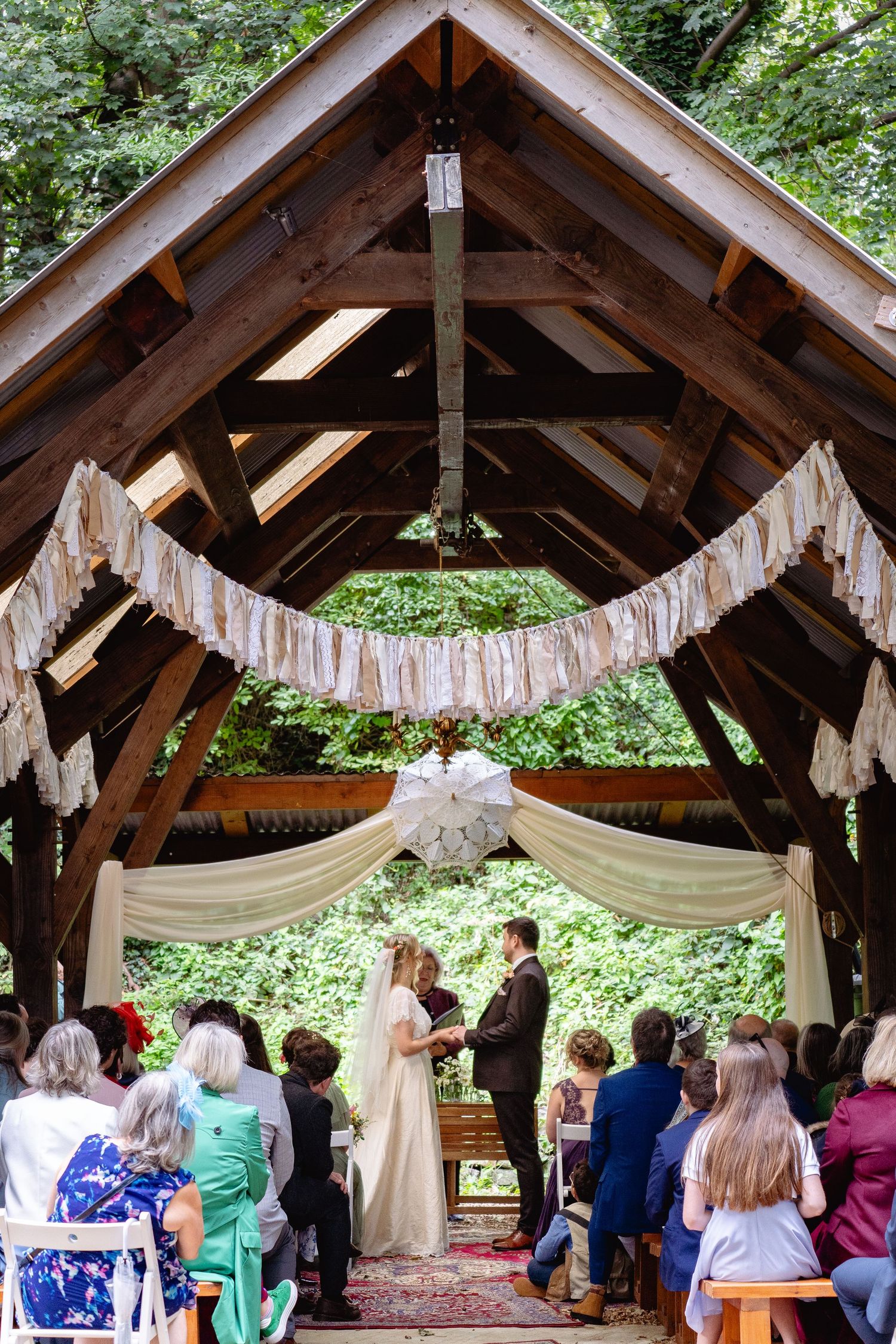 Wide shot of outdoor woodland wedding ceremony under rustic wooden pavilion with fabric bunting and seated guests.