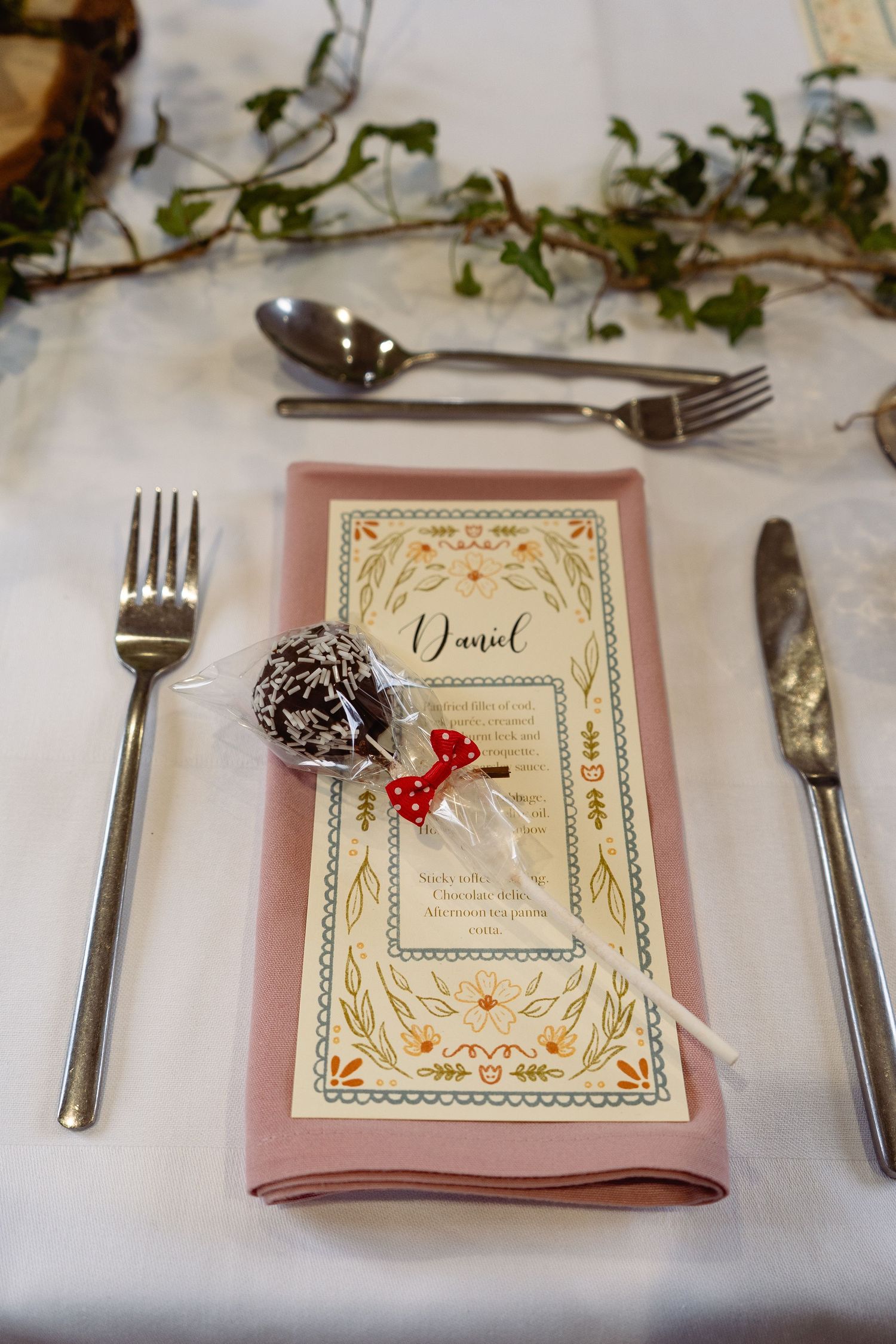 Elegant wedding place setting with pink napkin, menu card named Daniel, chocolate cake pop, and silver cutlery on white tablecloth.
