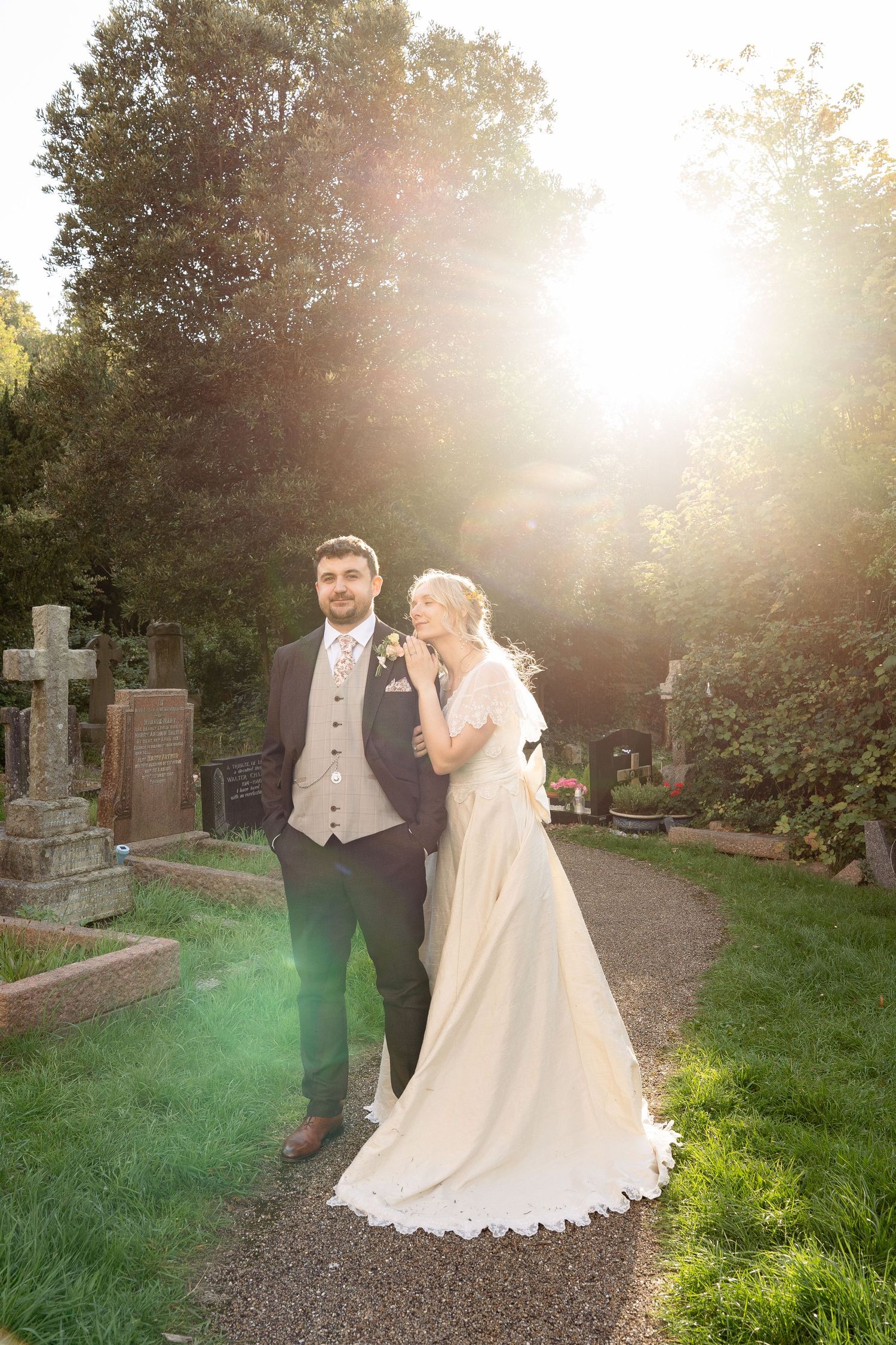 Bride and groom pose on a sunlit garden path surrounded by lush greenery and stone monuments on their wedding day.
