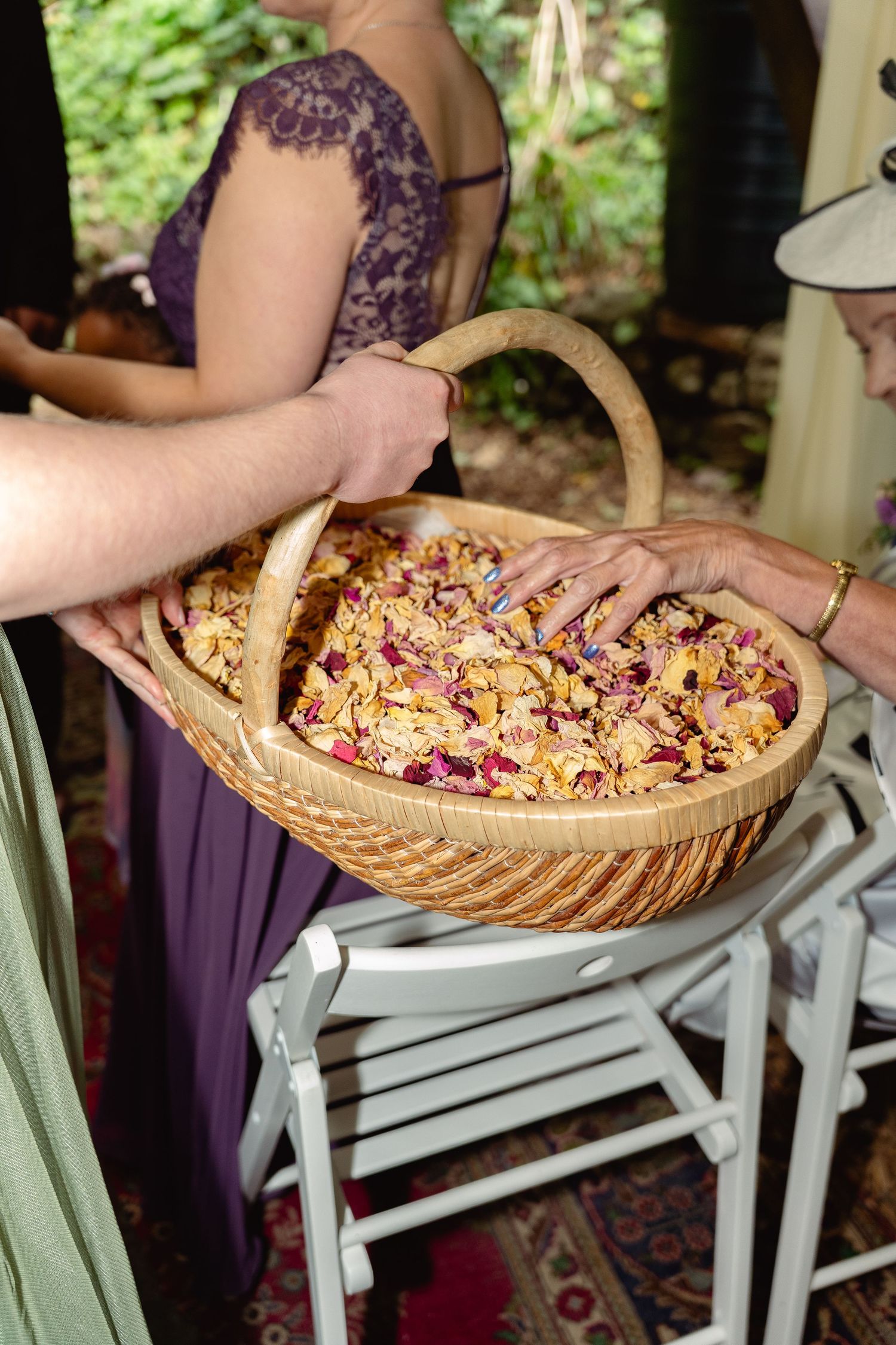 Hands reach into a wicker basket filled with colorful rose petals at an outdoor wedding ceremony.