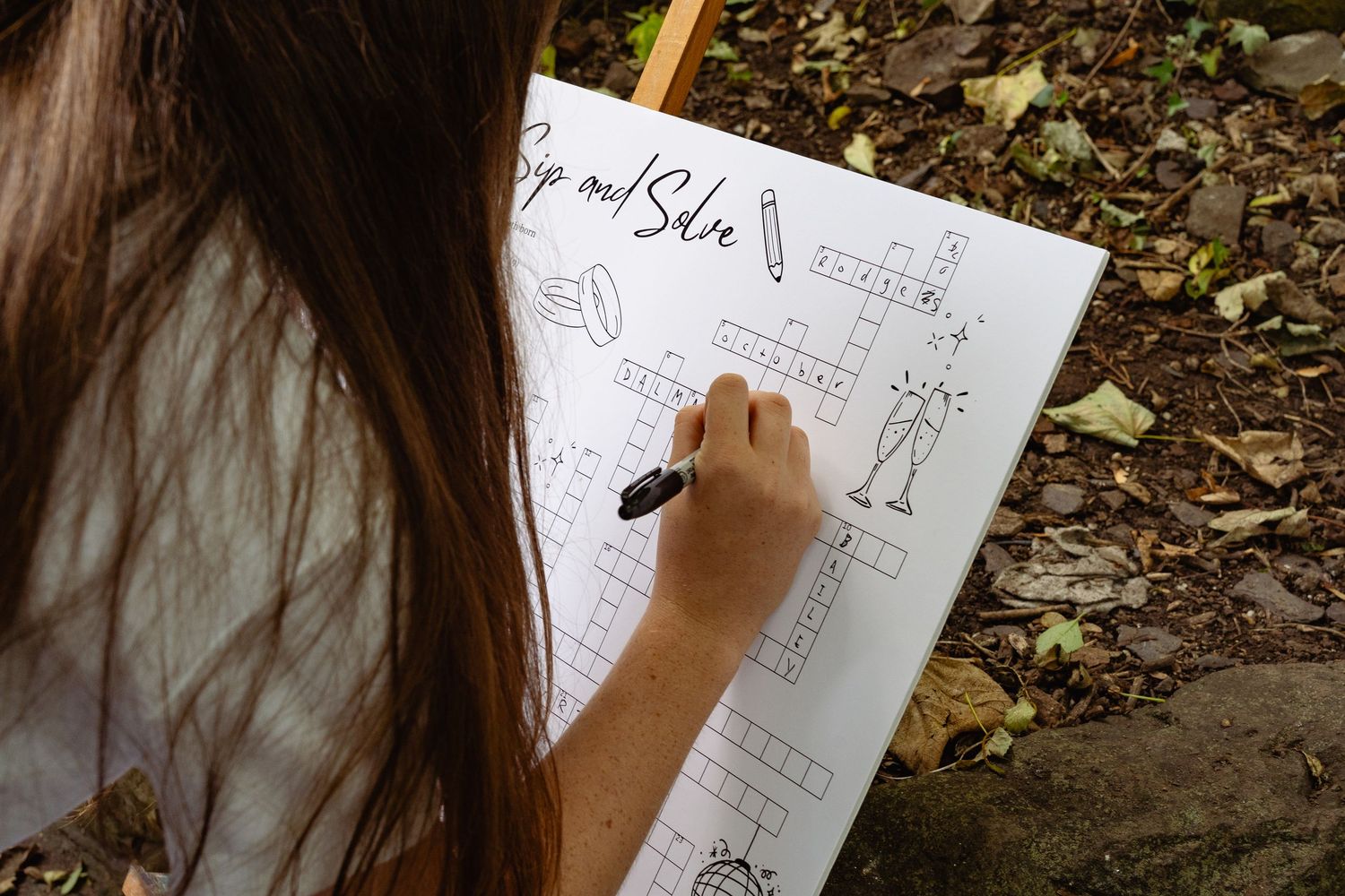 Woman with long brown hair writing on a crossword puzzle sign with a black marker at an outdoor wedding.
