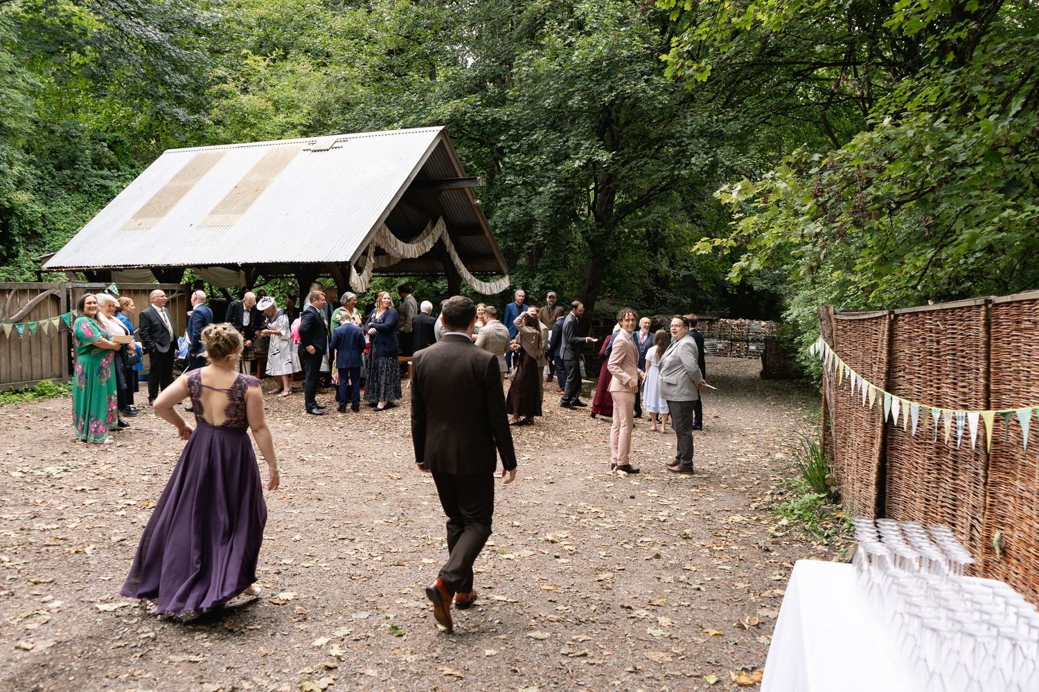 Guests gather outside a rustic woodland wedding venue with bunting decorations and a corrugated iron roof shelter.