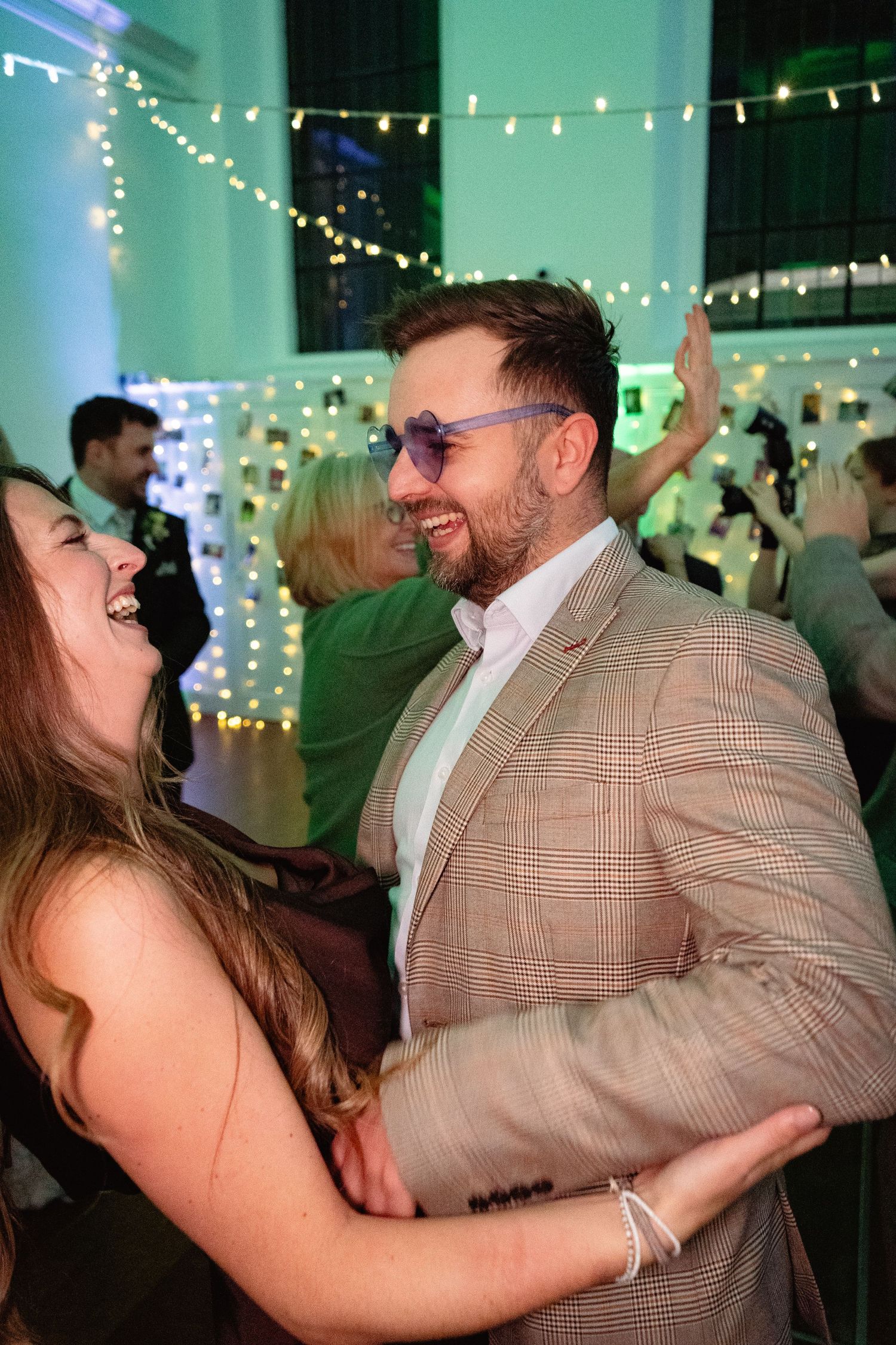 A laughing couple dances together at a wedding reception, surrounded by warm fairy lights, as the man wears stylish purple sunglasses.