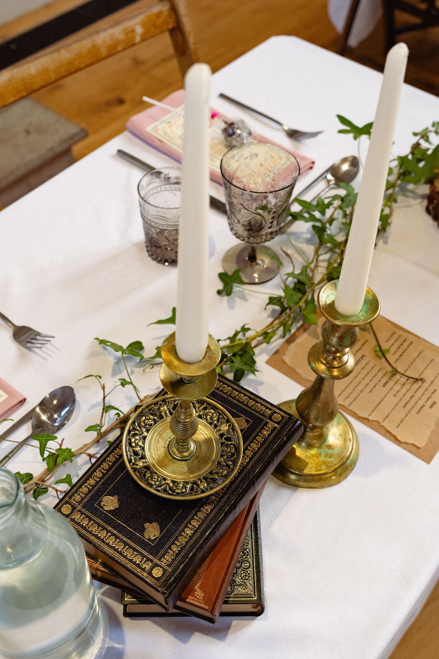 Elegant table setting with brass candlesticks, white taper candles, stacked books, ivy garland, and vintage glassware on white linen.