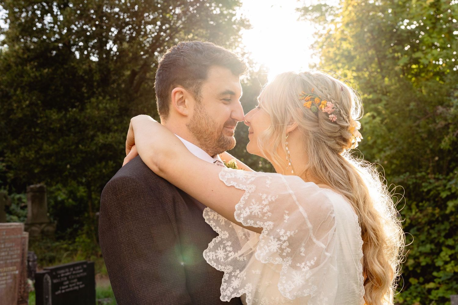 Bride and groom share a romantic moment outdoors, bathed in golden sunlight, with the bride wearing a lace dress and floral hair accessory.