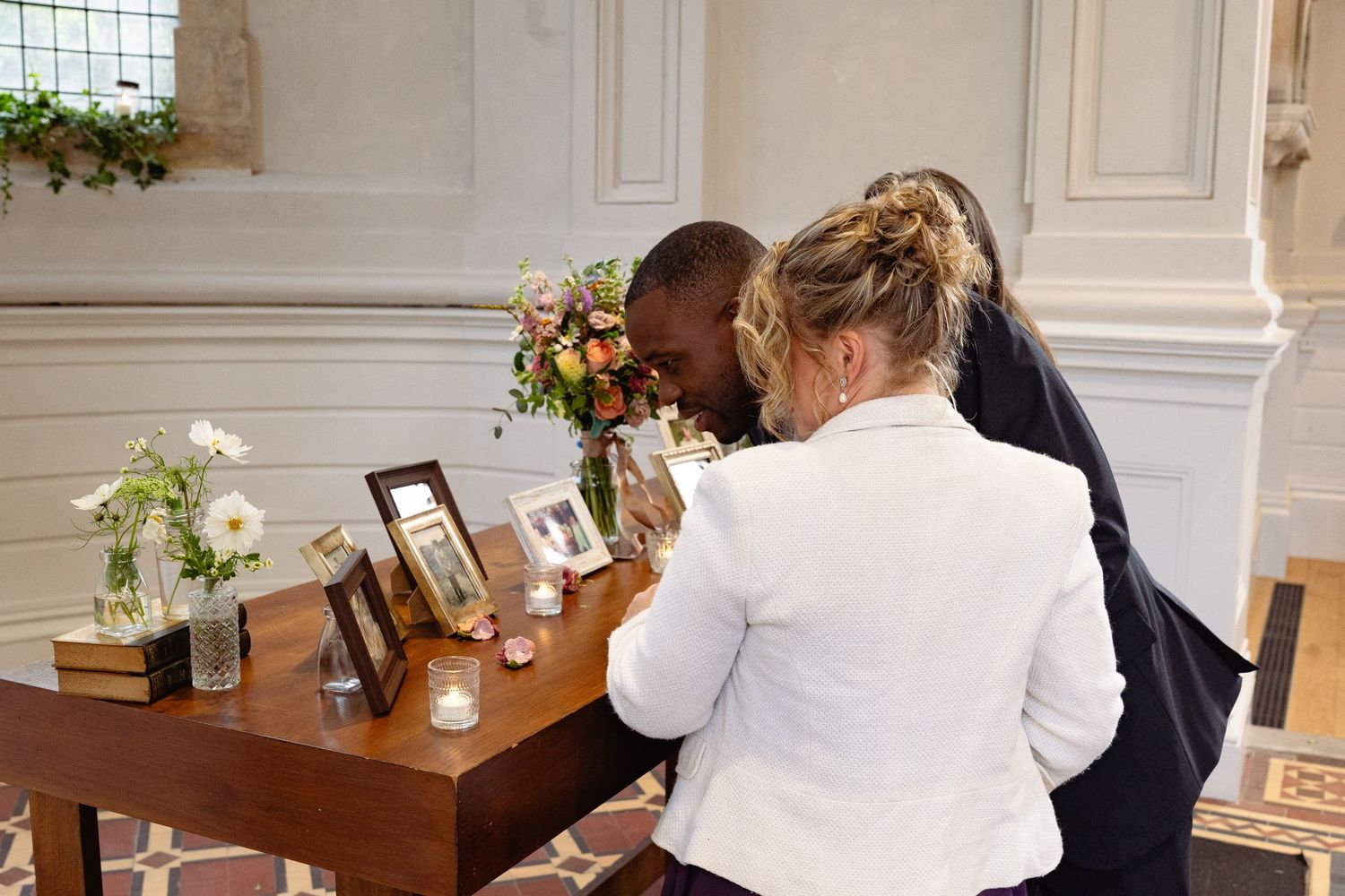 Couple signing wedding register at decorated table with flowers and framed photos in elegant venue.