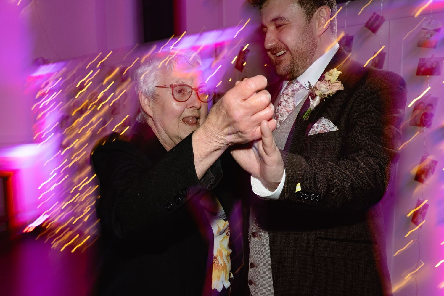 Elderly woman and groom share a joyful dance at a wedding reception with colorful purple lighting.
