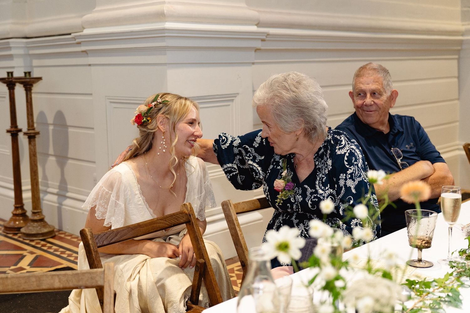Bride in floral crown laughs with elderly woman at elegant wedding reception table with white flowers.