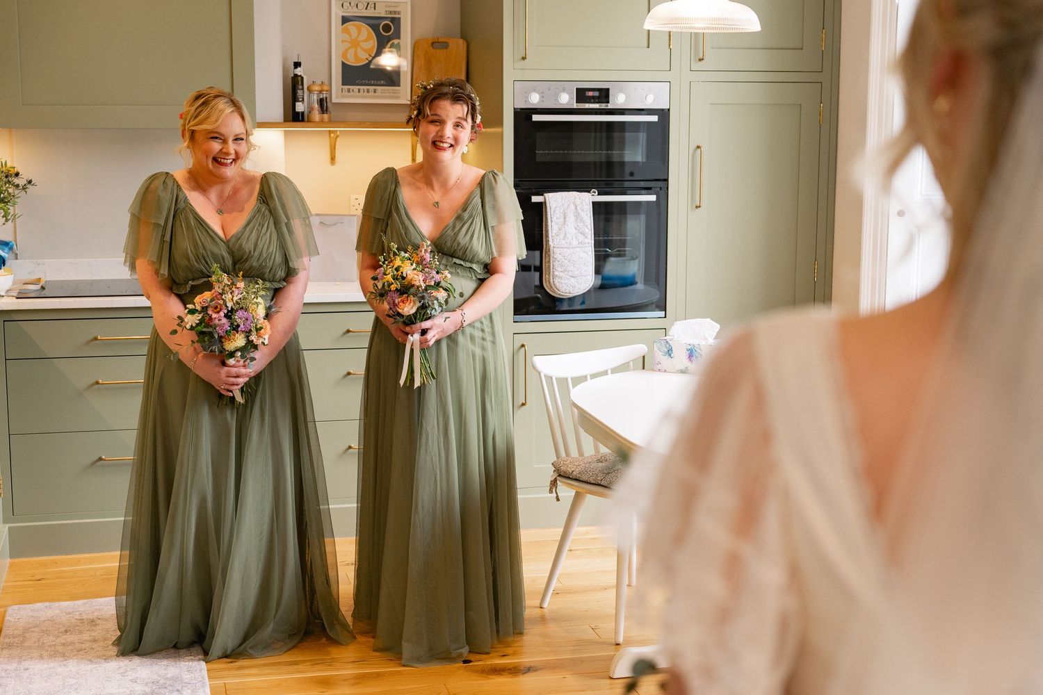 Two bridesmaids in sage green dresses holding bouquets smile at bride in white gown in elegant kitchen setting.