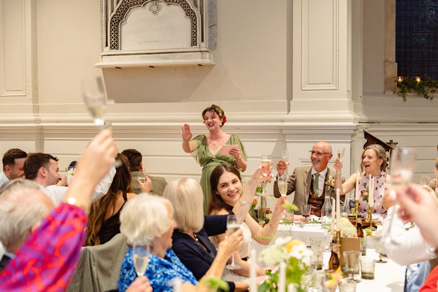 Wedding guests raising glasses in a toast during a reception in an elegant white hall.