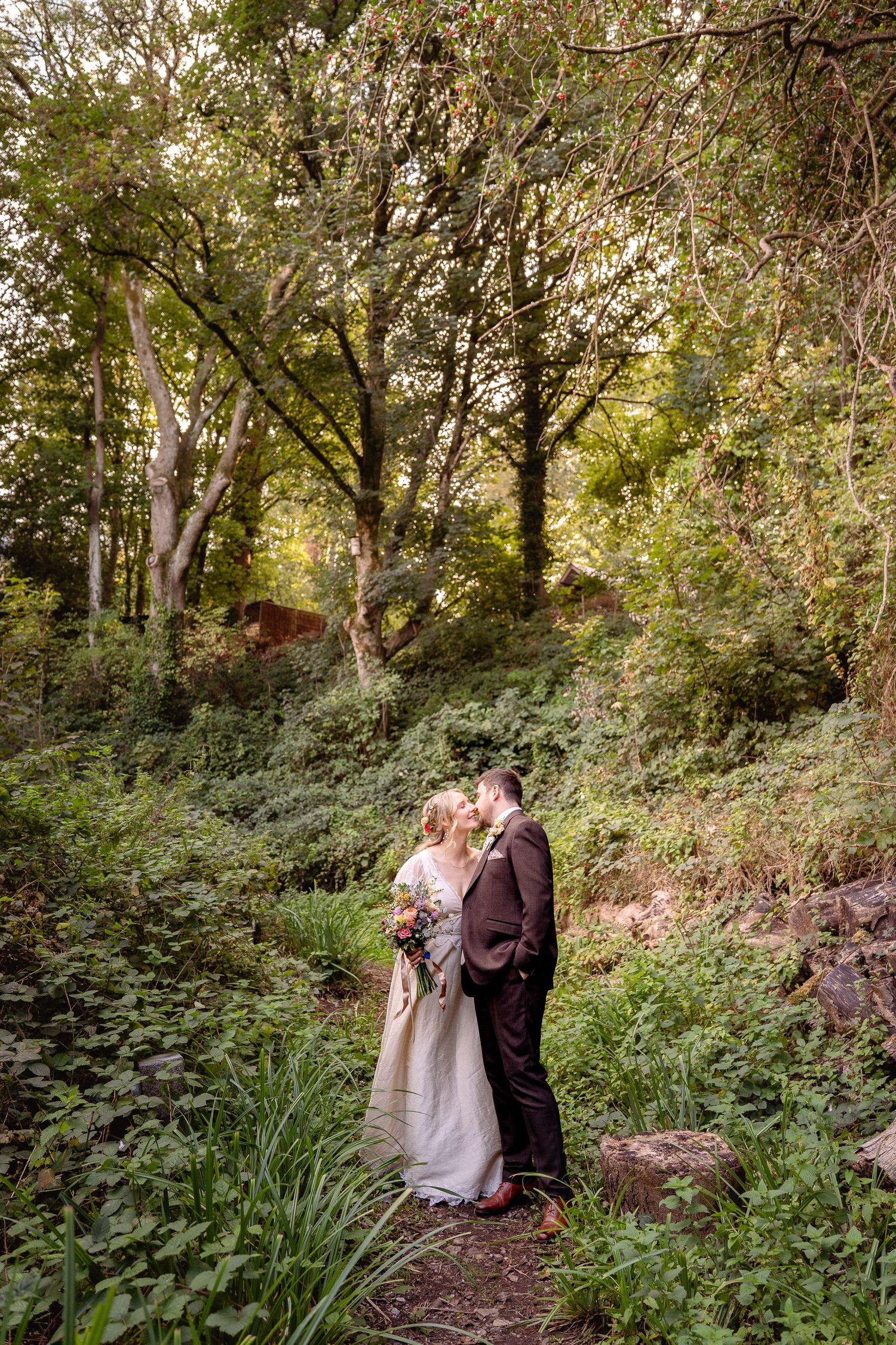 Bride and groom share a romantic kiss surrounded by lush green trees and foliage in a forest wedding portrait.