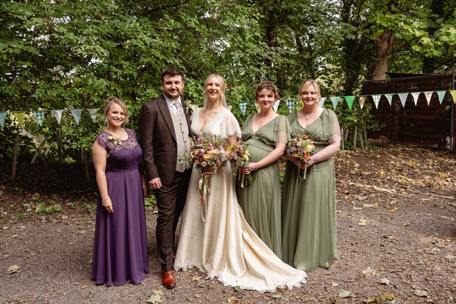 Wedding party posing outdoors with bride in ivory gown, groom, and bridesmaids in purple and sage green dresses.