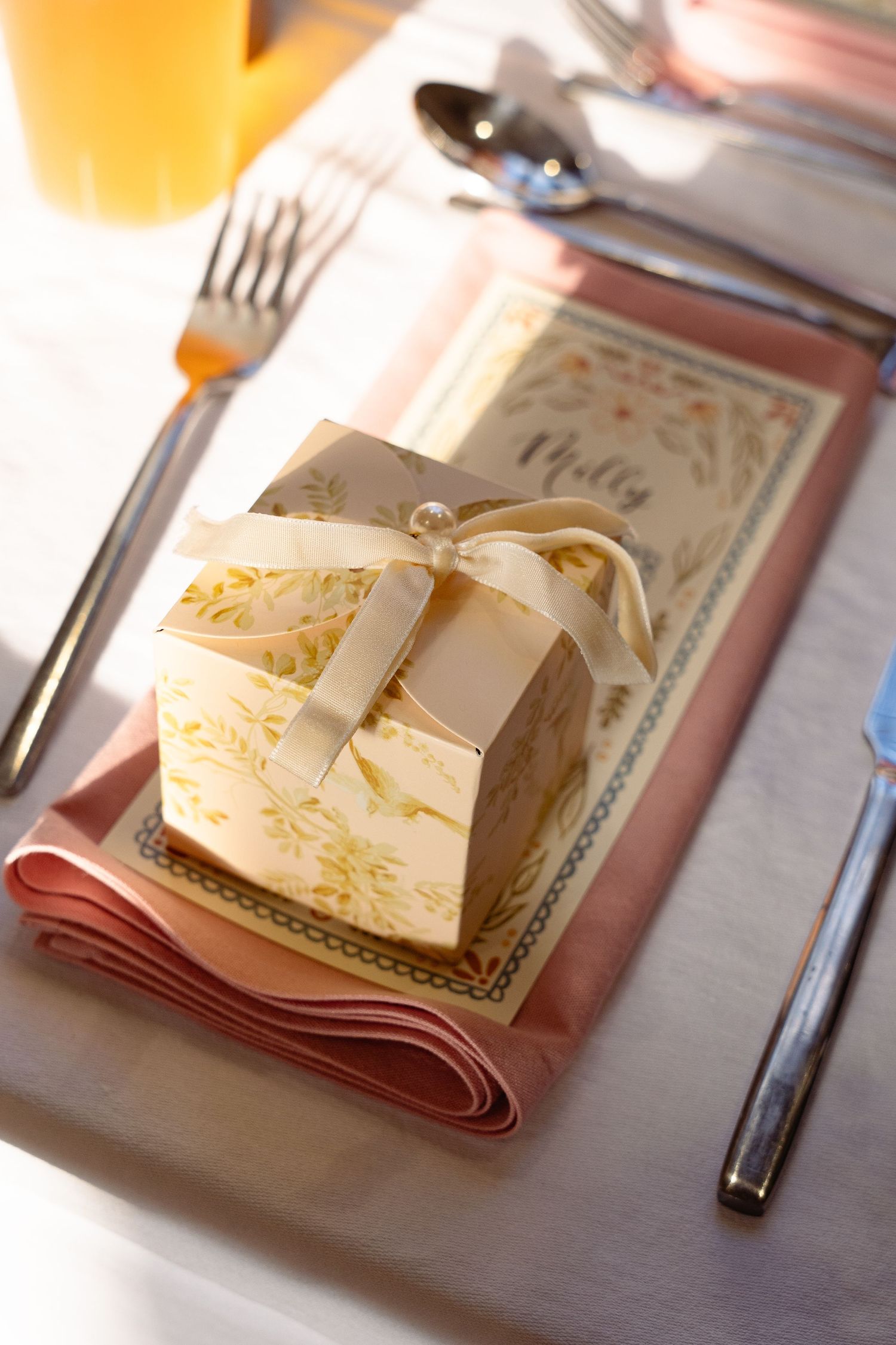 Elegant wedding favor box with floral design and pearl ribbon on a pink napkin beside silverware and place card.