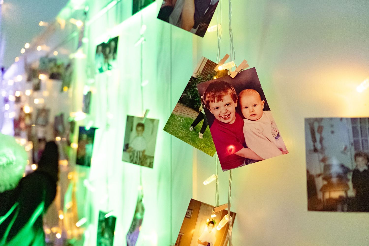 Photos of children displayed on a wall with fairy lights at a wedding reception.