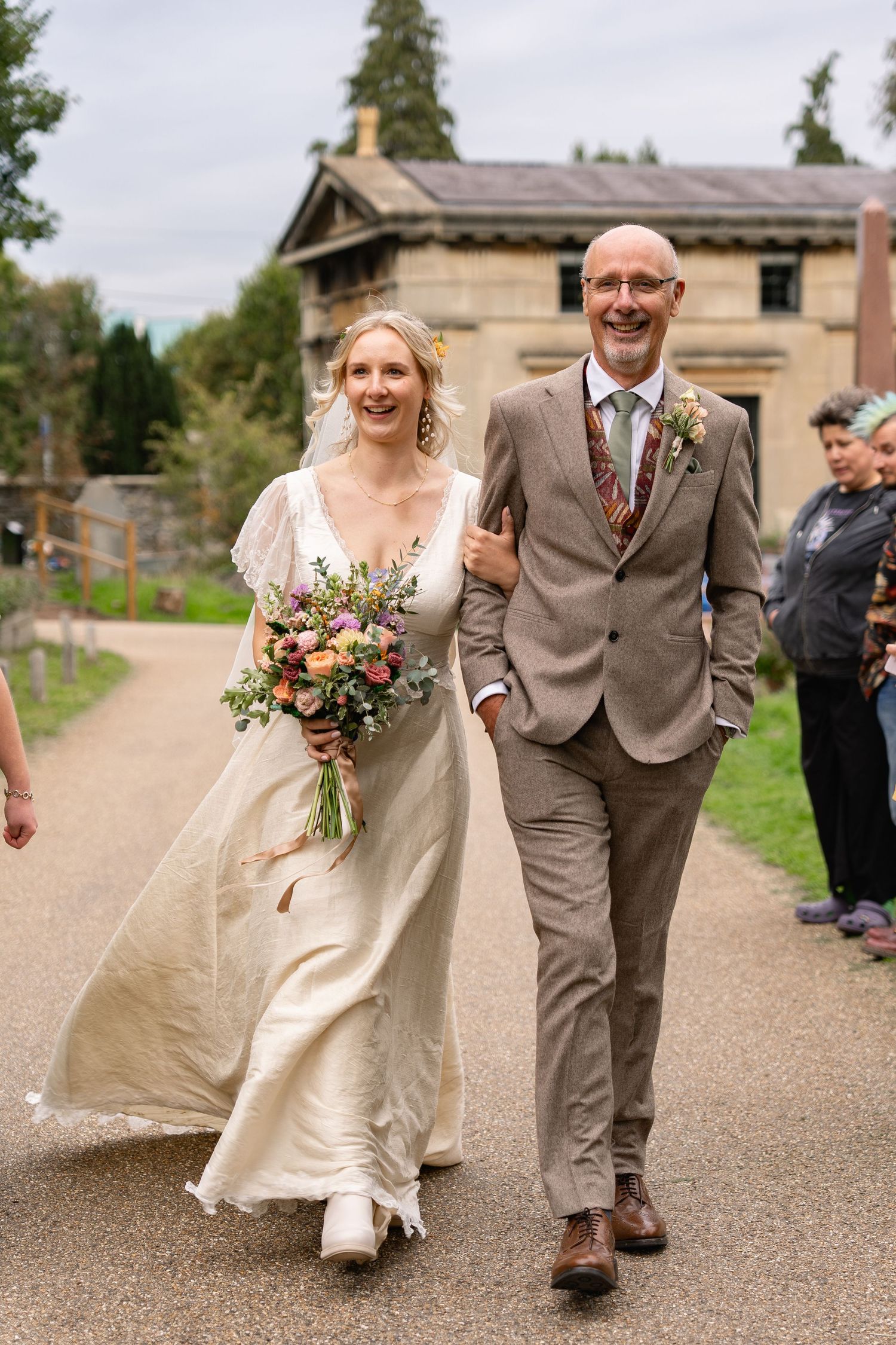 Smiling bride in white gown holding colorful bouquet walks with bald suited man at outdoor wedding venue.