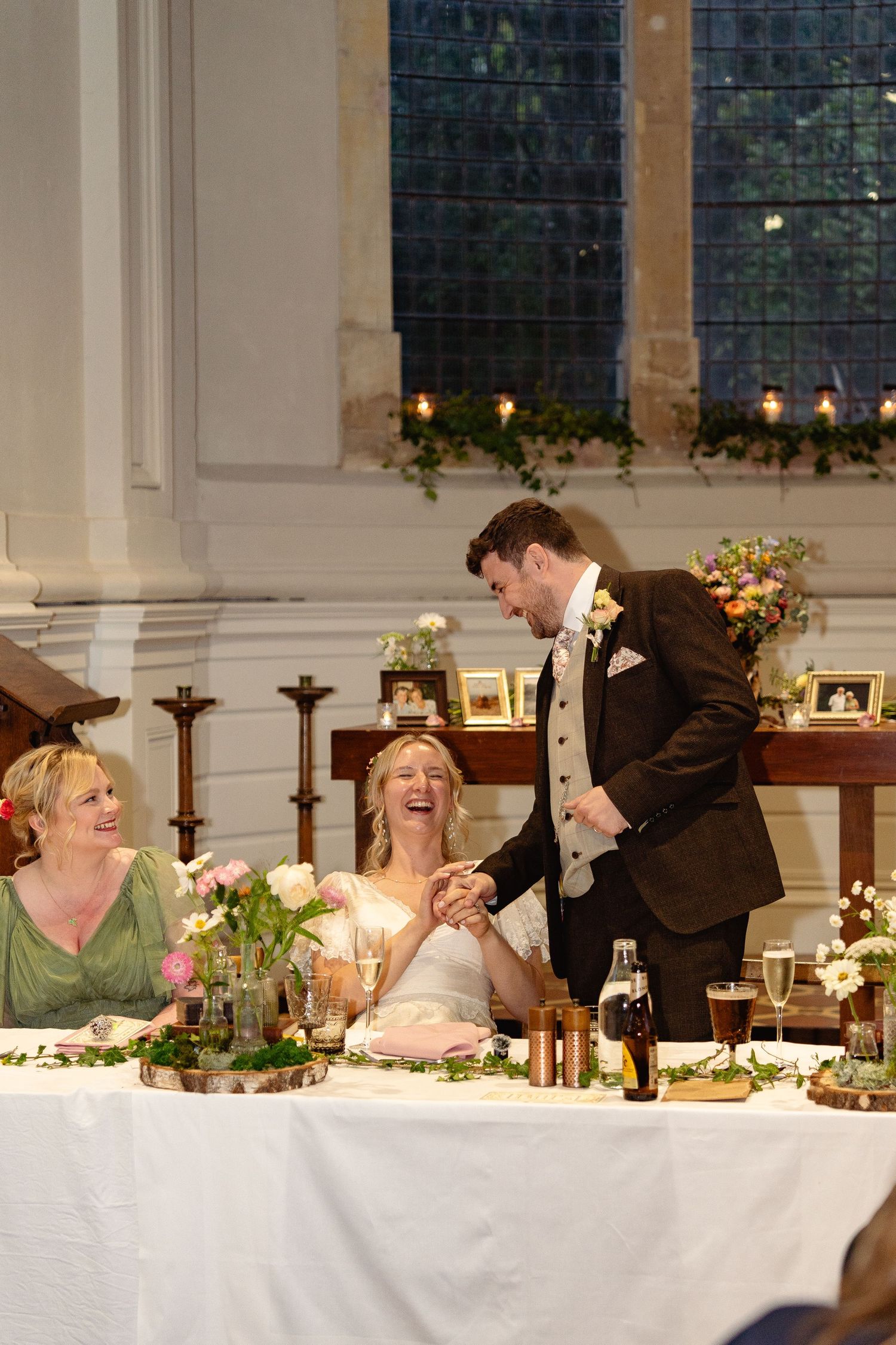 Bride and groom laughing together at wedding reception head table, with bridesmaid seated nearby in elegant church venue.