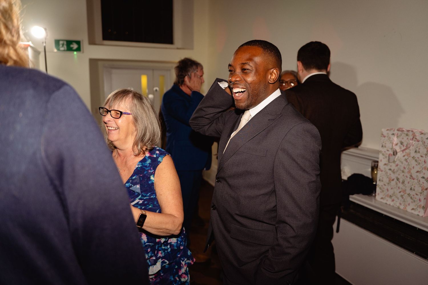 Guests socializing and laughing at a formal evening event, man in grey suit smiling with woman in blue dress.
