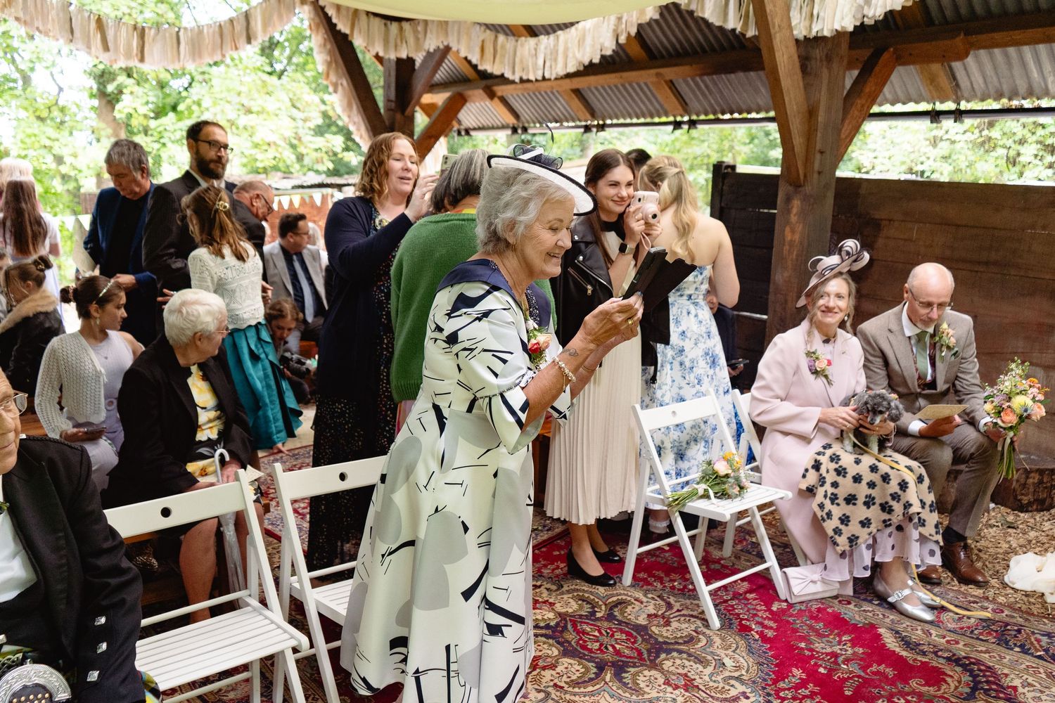 Guests gather at an outdoor wedding ceremony under a rustic wooden pavilion, seated on white chairs atop a colorful rug.
