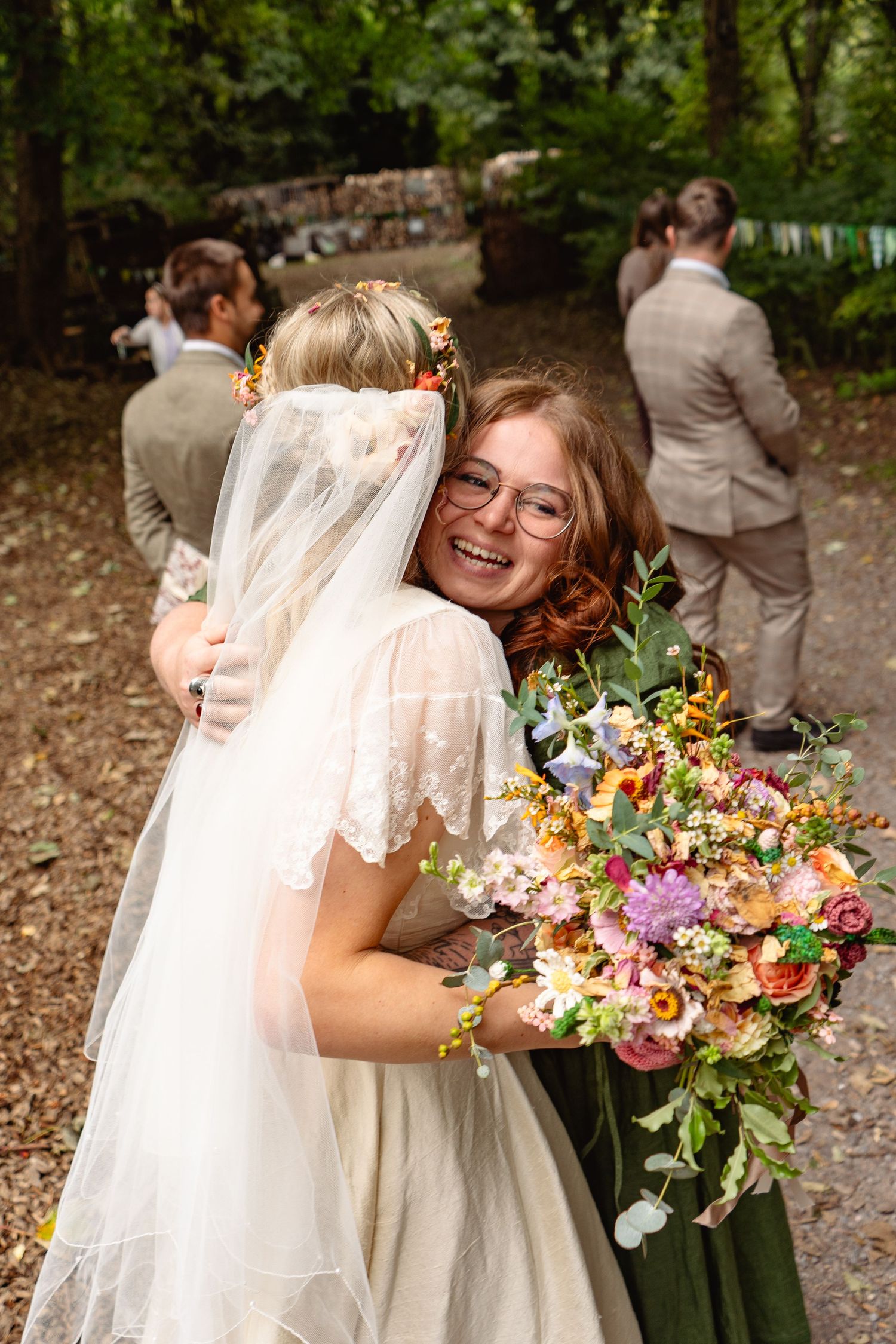 Smiling bridesmaid with glasses hugs bride in white wedding dress and veil, holding colorful bridal bouquet outdoors.