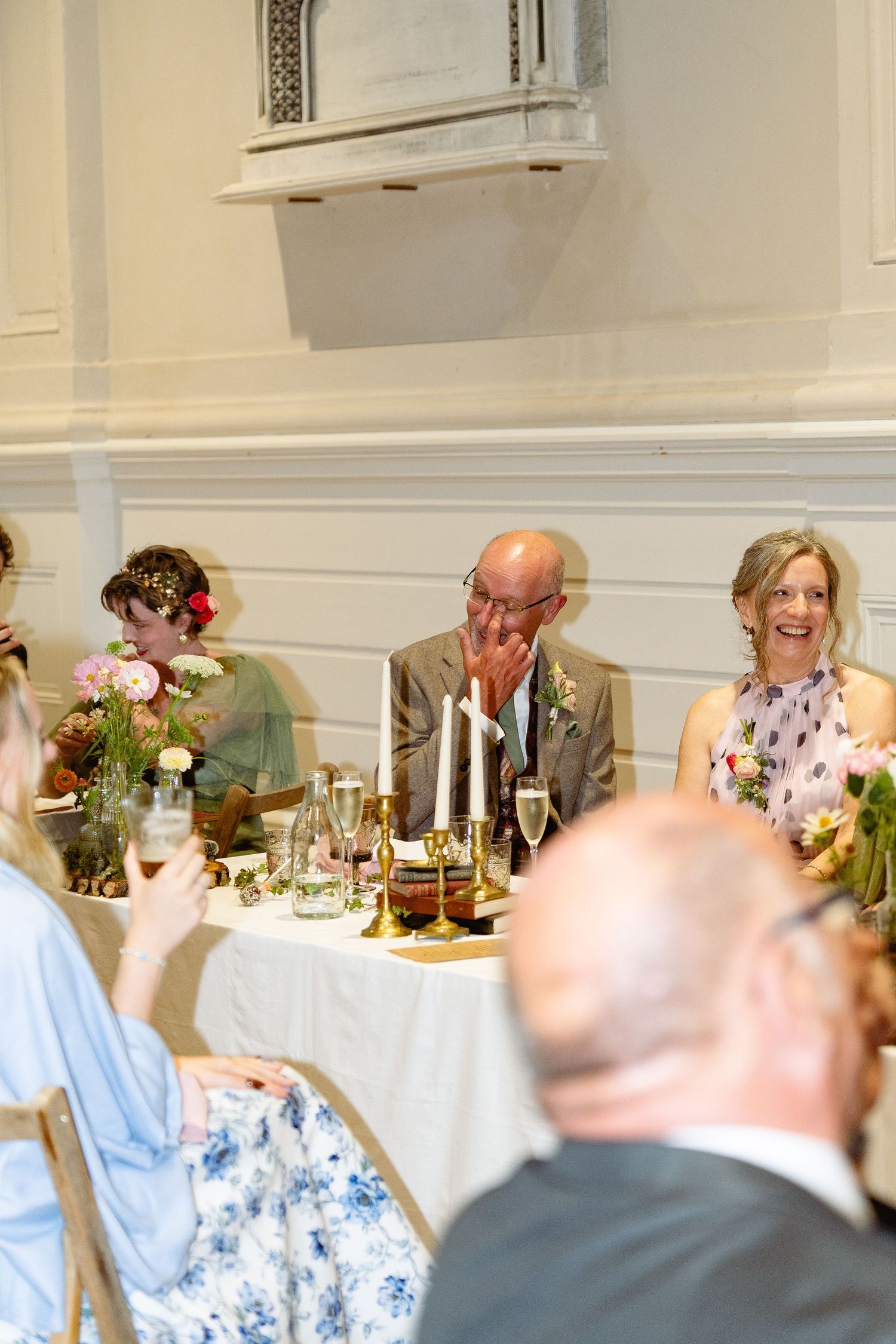 Guests laughing and celebrating at a wedding reception dinner table with floral arrangements and candles.