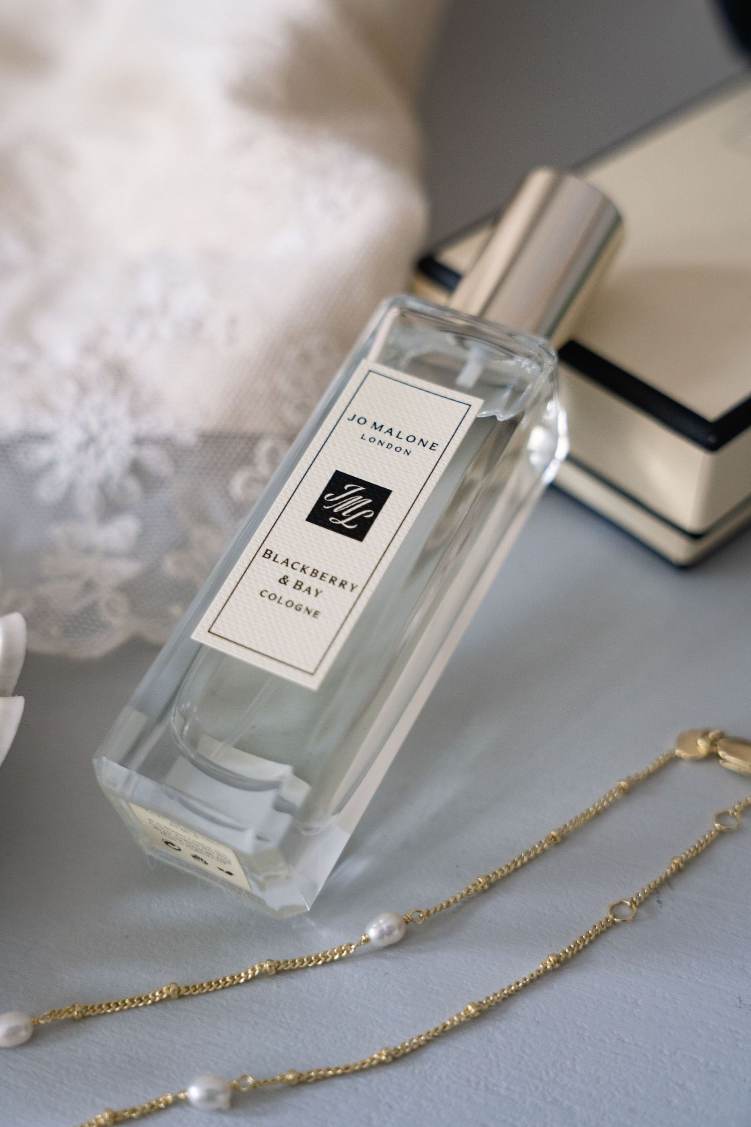 Jo Malone London Blackberry & Bay Cologne bottle beside a pearl necklace and lace fabric on a soft blue surface.
