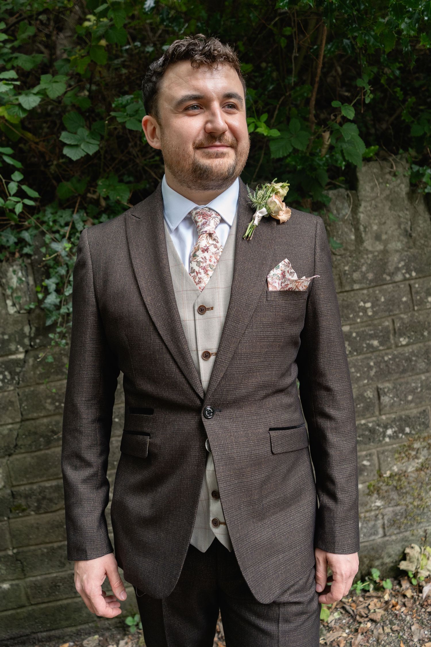 Groom in brown tweed suit with floral tie, pocket square, and boutonniere posing outdoors against a stone wall.