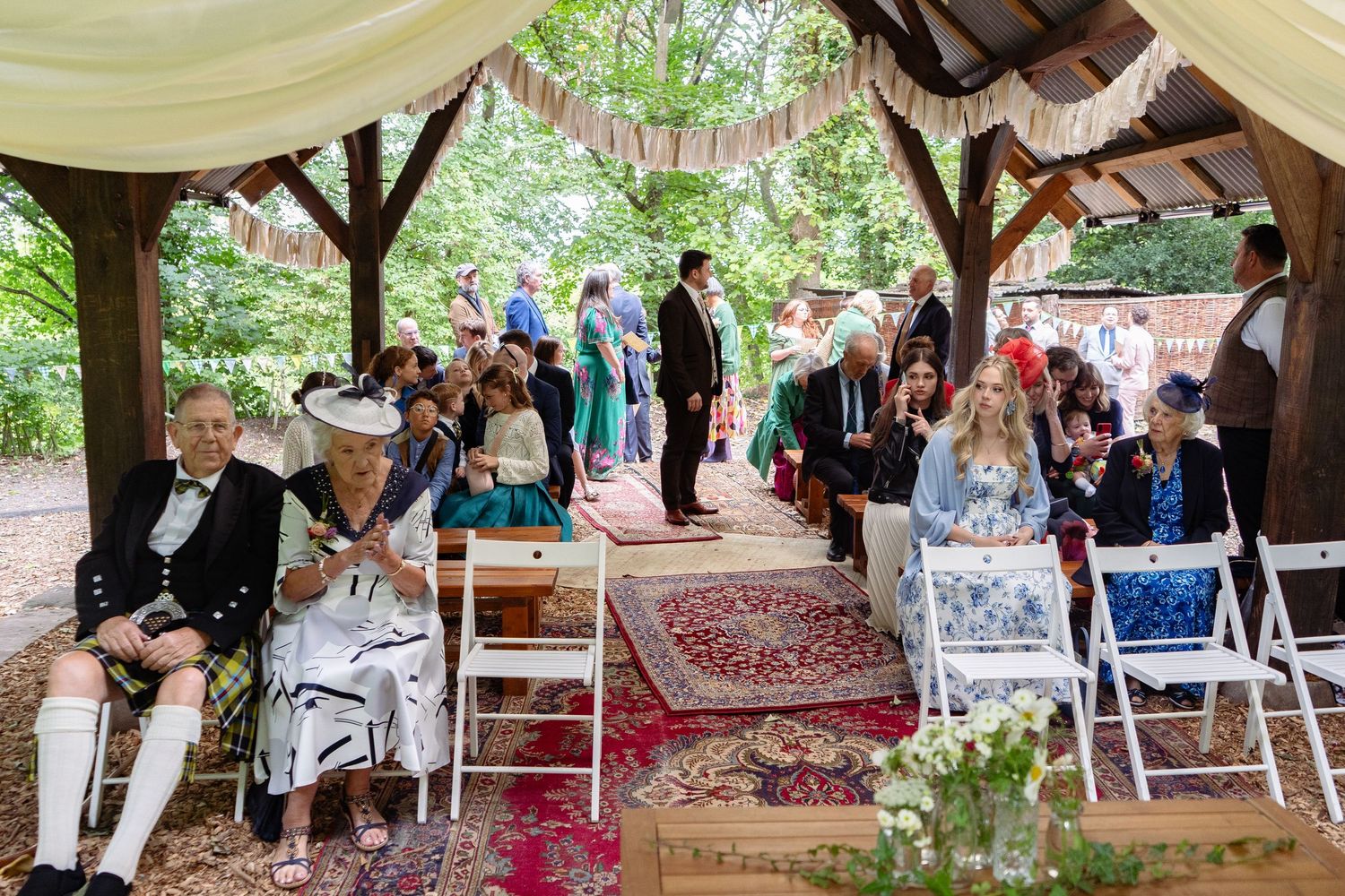 Guests seated at an outdoor woodland wedding ceremony under a rustic wooden gazebo decorated with white draping.