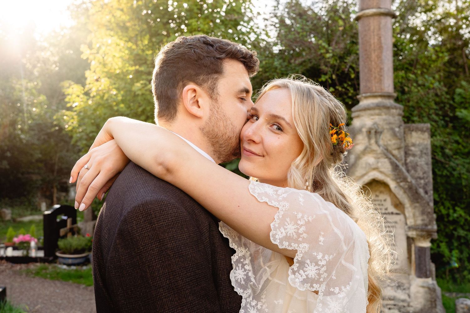 A bride and groom embrace tenderly in a sunlit garden, the bride in lace smiling softly as the groom nuzzles her cheek.