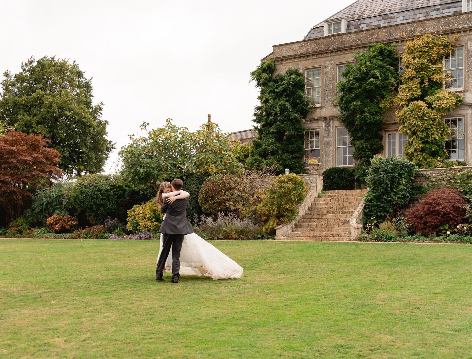 Bride and groom embrace on lush green lawn of Hamswell House in Bath before the elegant stone manor house with manicured gardens.