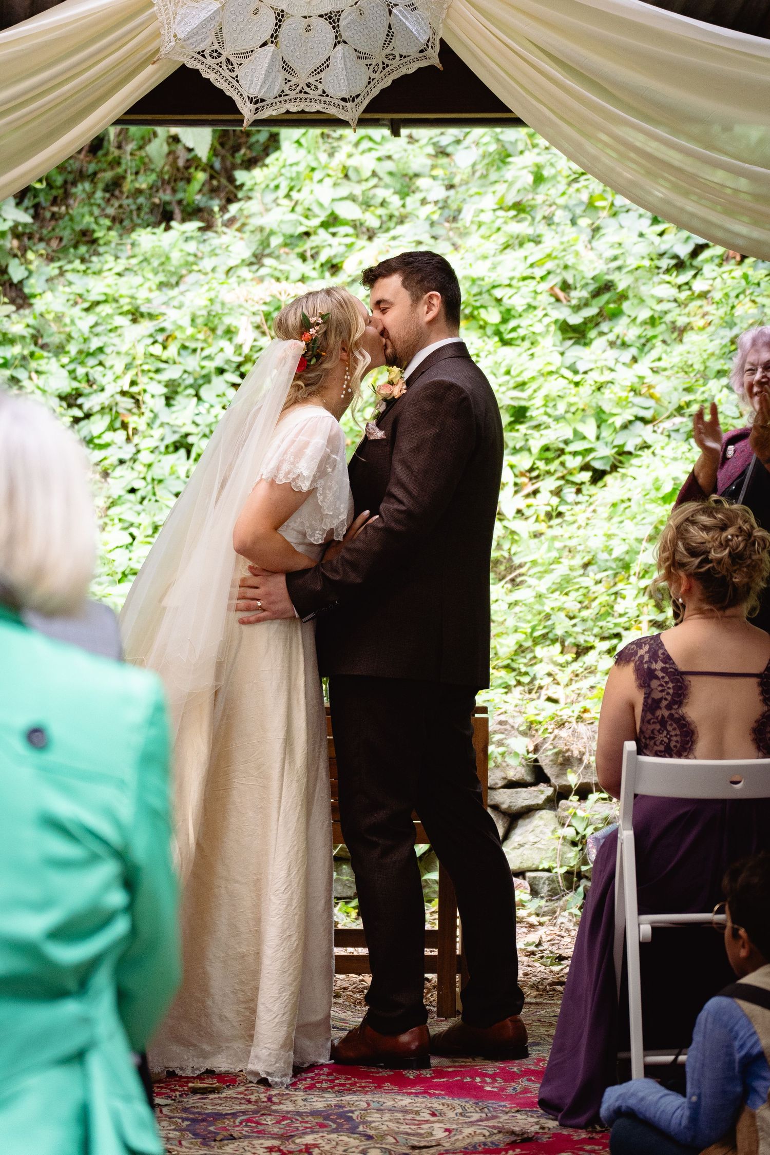 Bride and groom share first kiss at outdoor woodland wedding ceremony beneath white draped canopy.