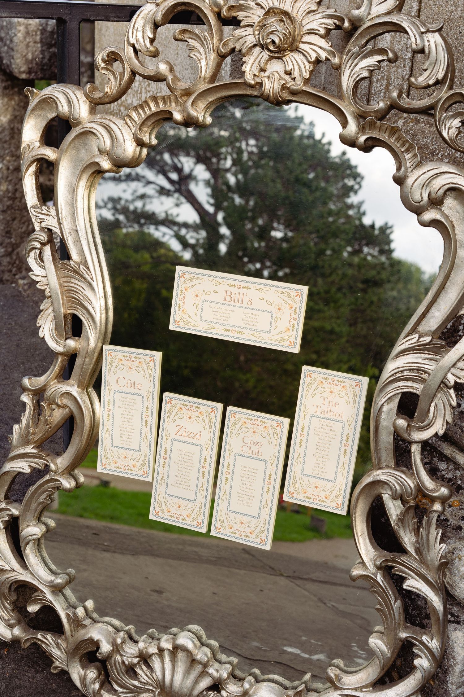 Ornate silver baroque mirror displaying elegant wedding seating chart cards reflecting a lush green garden backdrop.