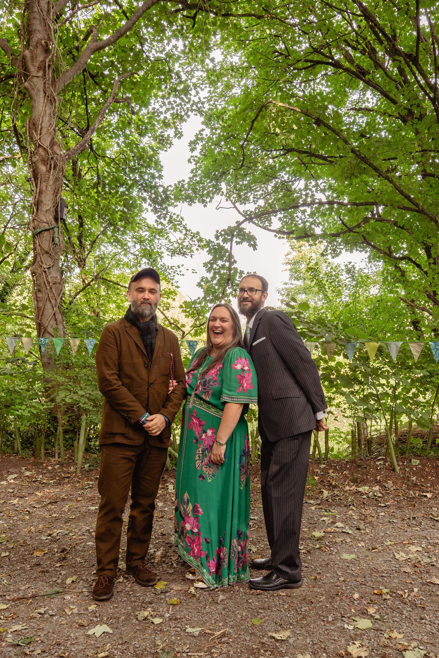 Three people posing outdoors in a woodland setting with bunting, two men in suits flanking a woman in a green floral dress.