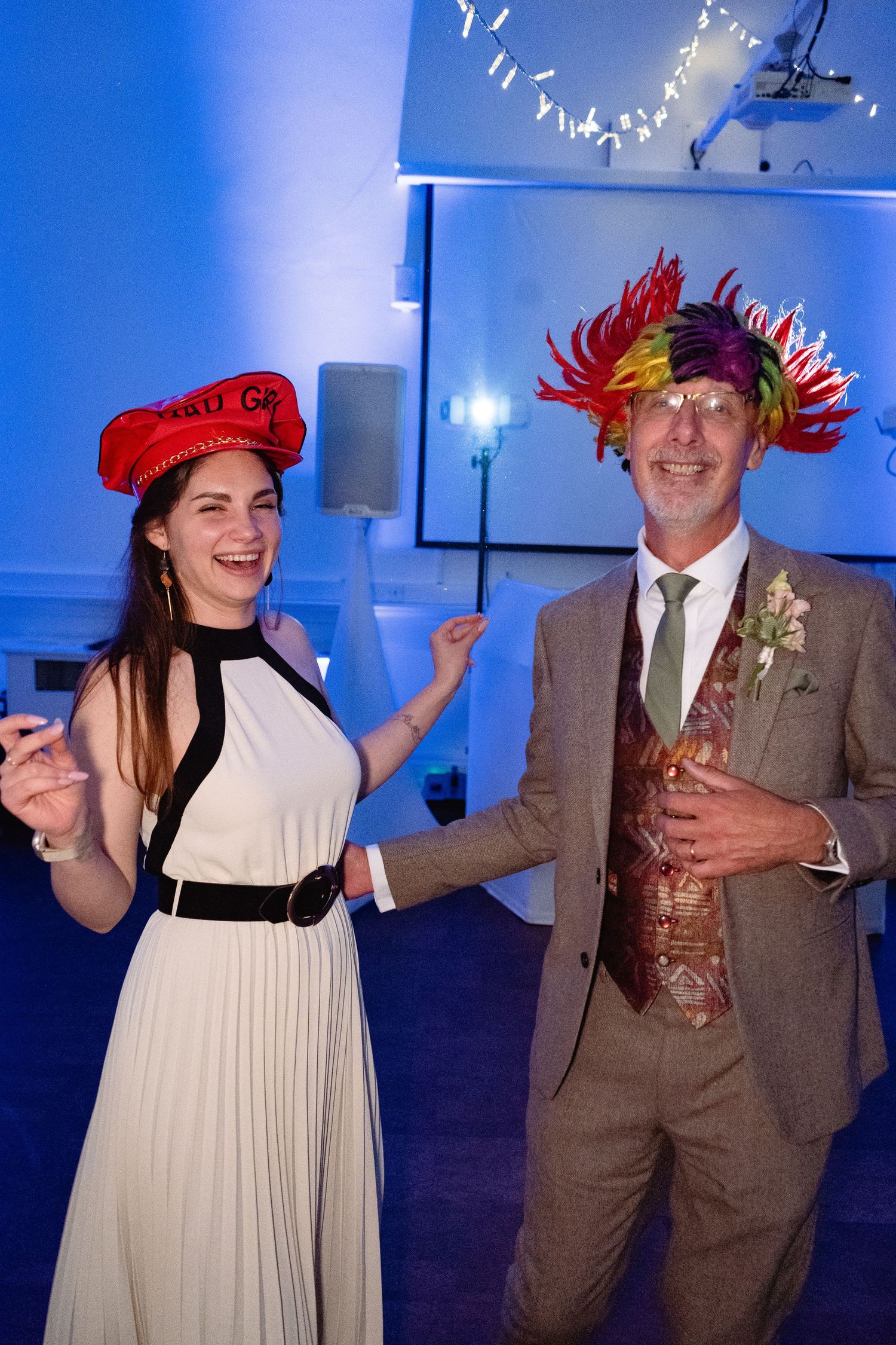 Two people wearing fun party hats dance together at a wedding reception with blue ambient lighting.