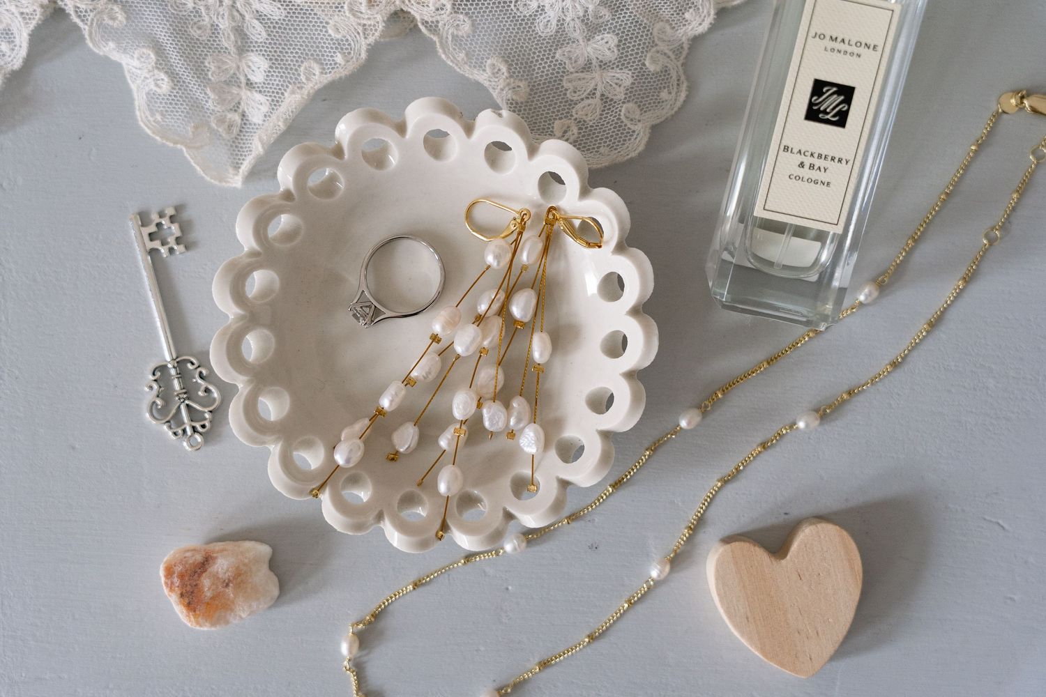 Flat lay of white scalloped jewelry dish with ring and earrings, Jo Malone perfume, heart stone, crystal, and delicate pearl necklace.