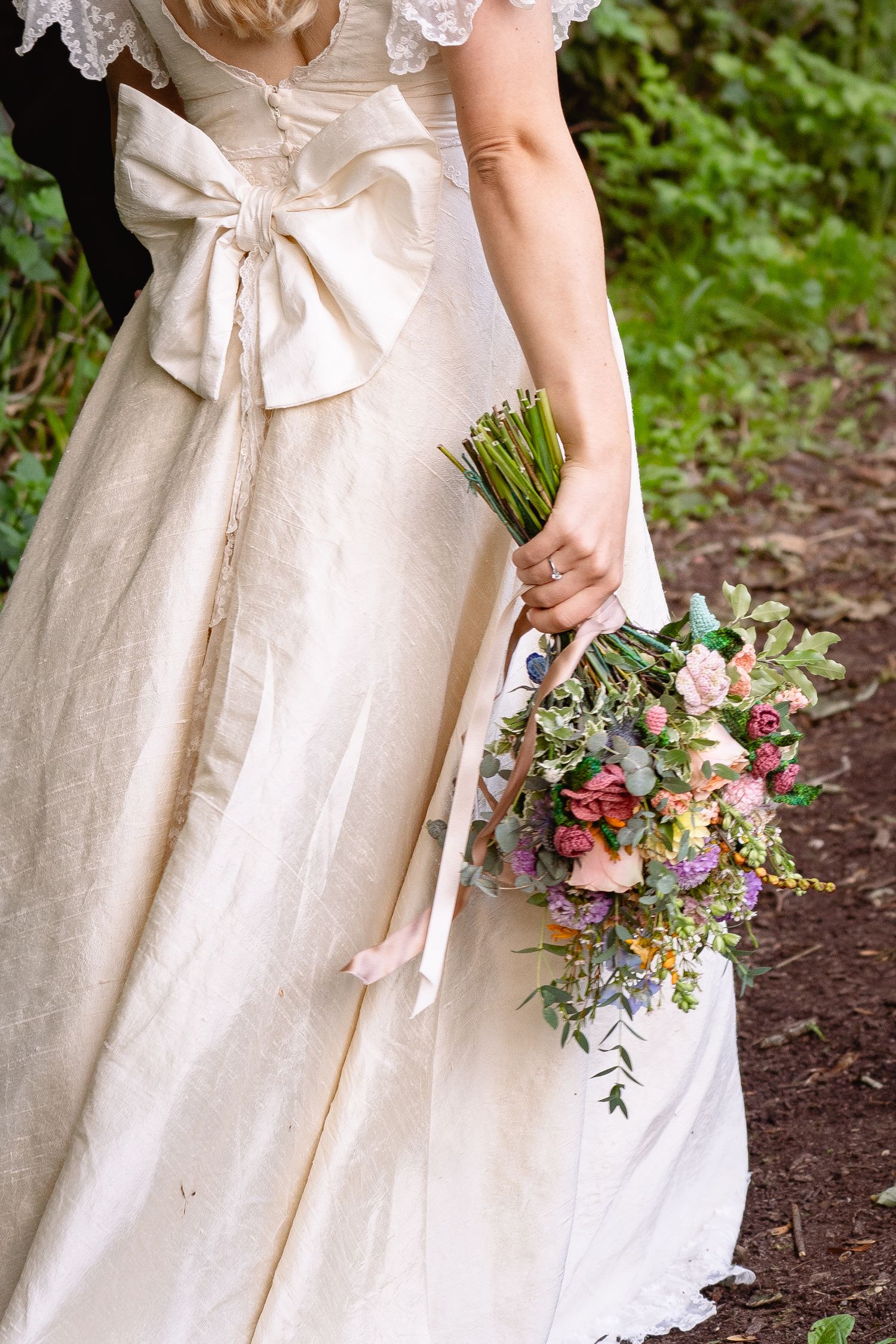 Bride in elegant ivory gown with large bow holding a colorful wildflower bouquet at an outdoor wedding.