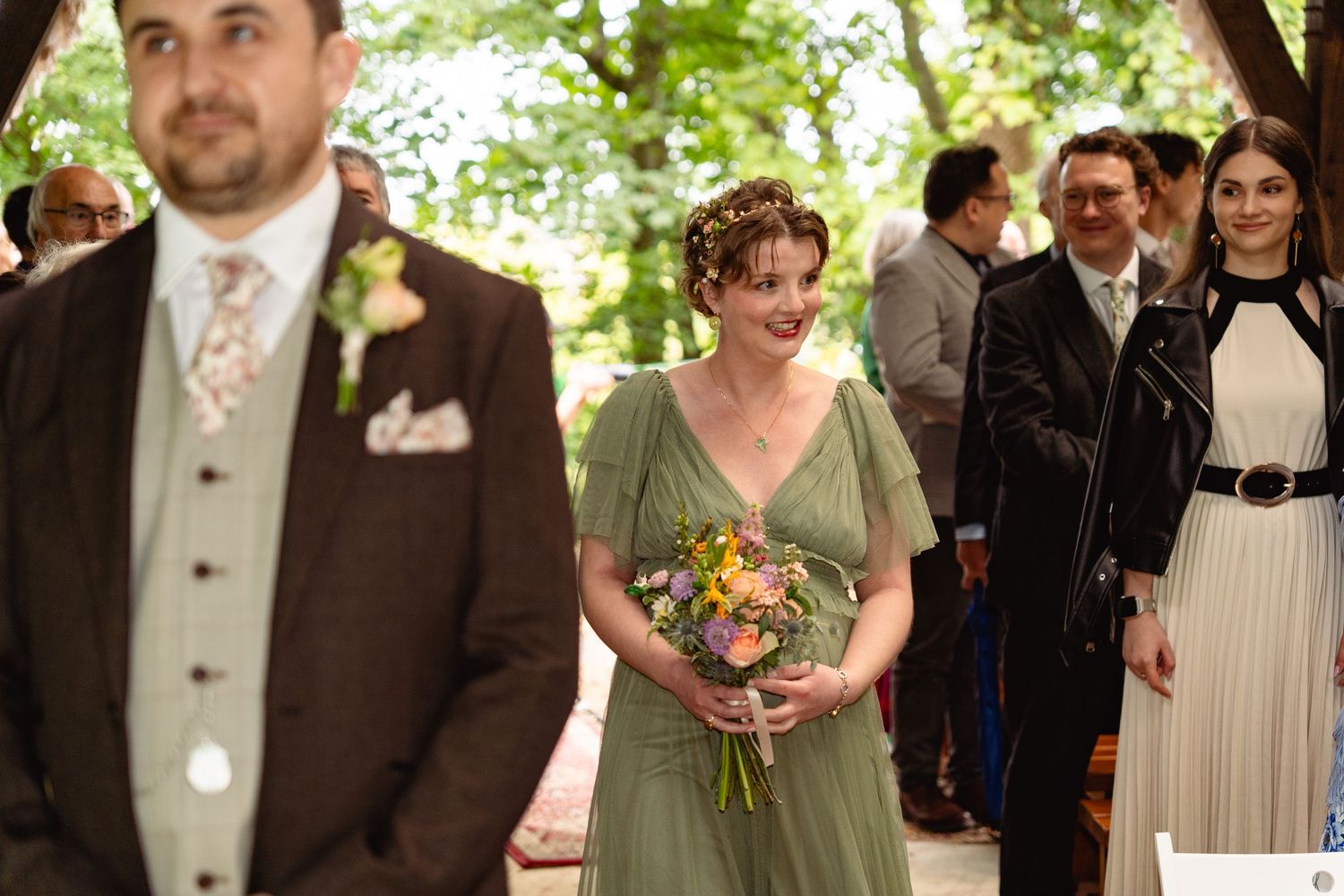 Smiling bridesmaid in sage green dress holding colorful bouquet walking down the aisle at wedding.