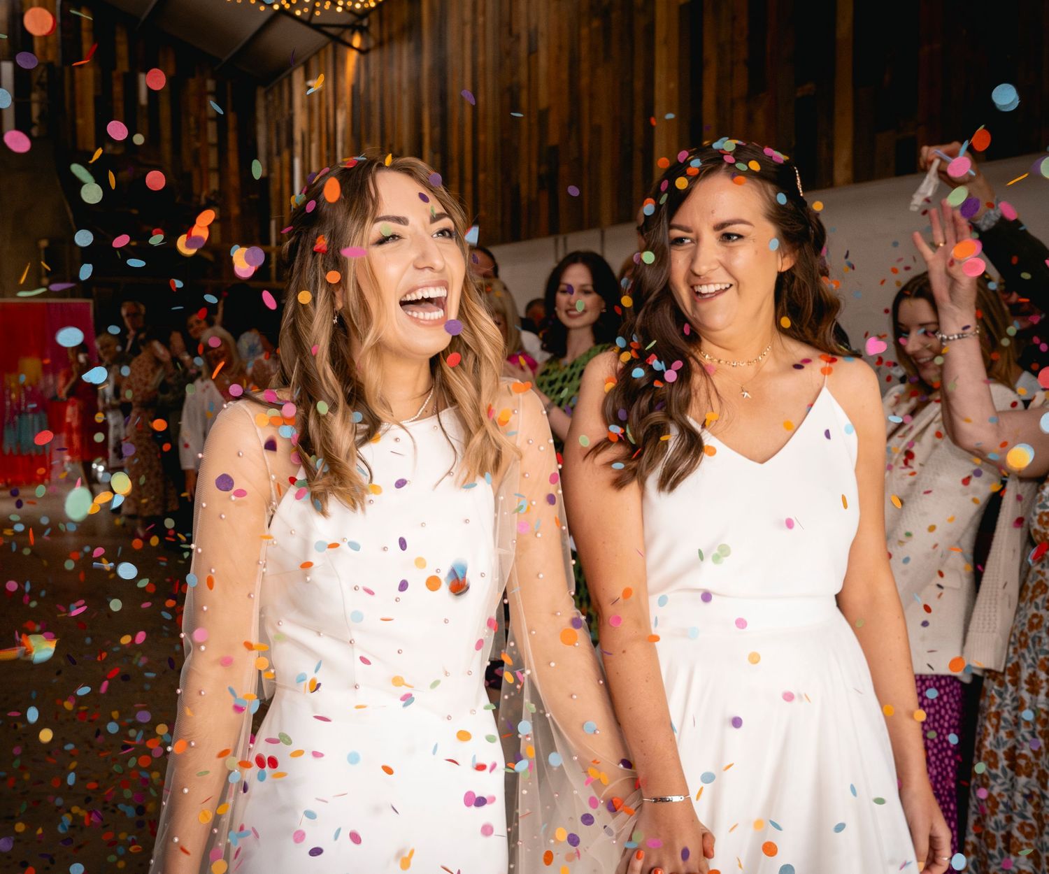 Two brides in white dresses laugh joyfully as colourful confetti rains down during their barn wedding celebration.
