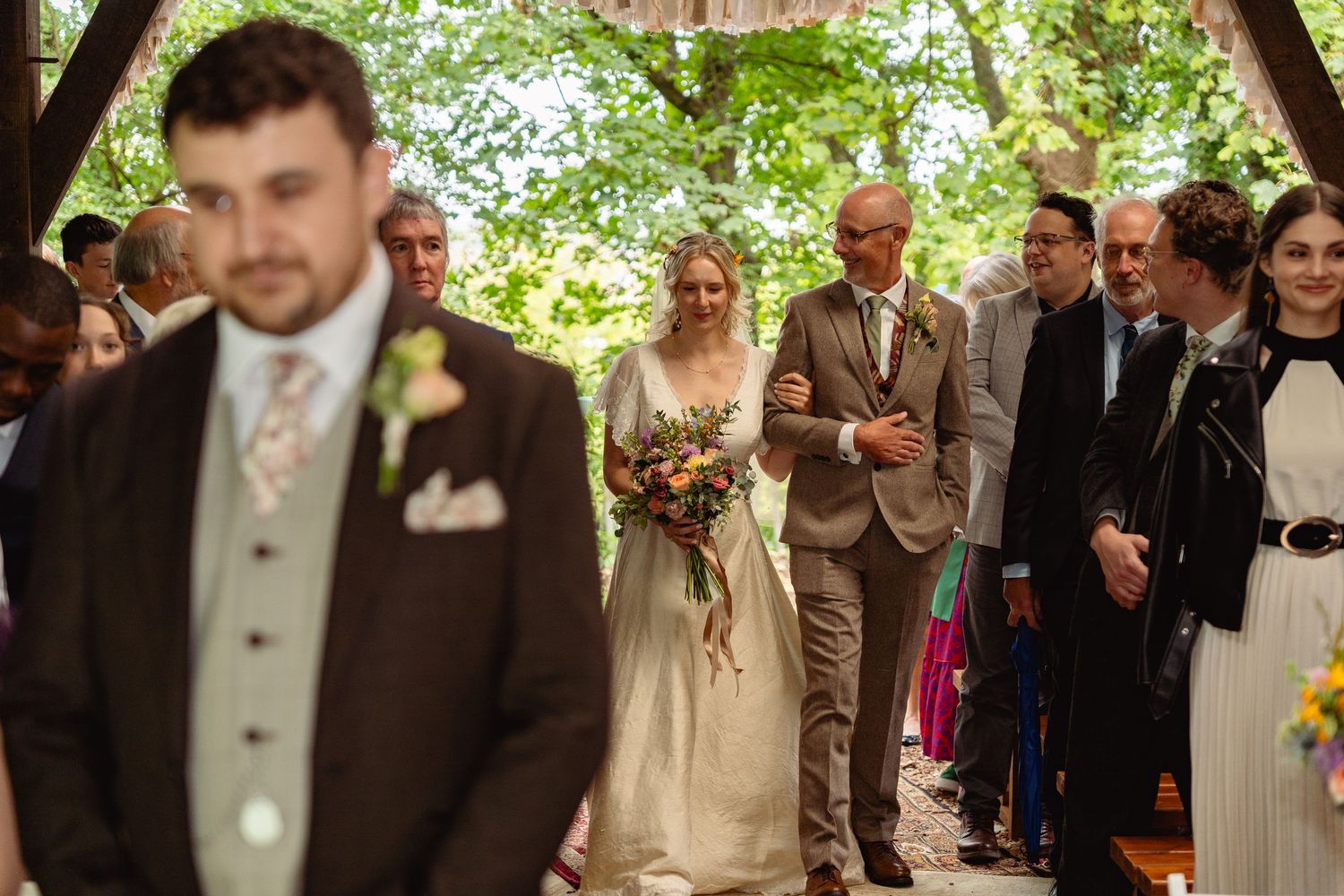 Bride in white gown walks down aisle with escort as groom waits in foreground at outdoor woodland wedding.
