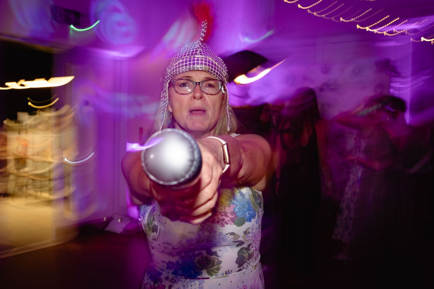 Woman in floral dress and headband holds glowing silver orb at a purple-lit party event.